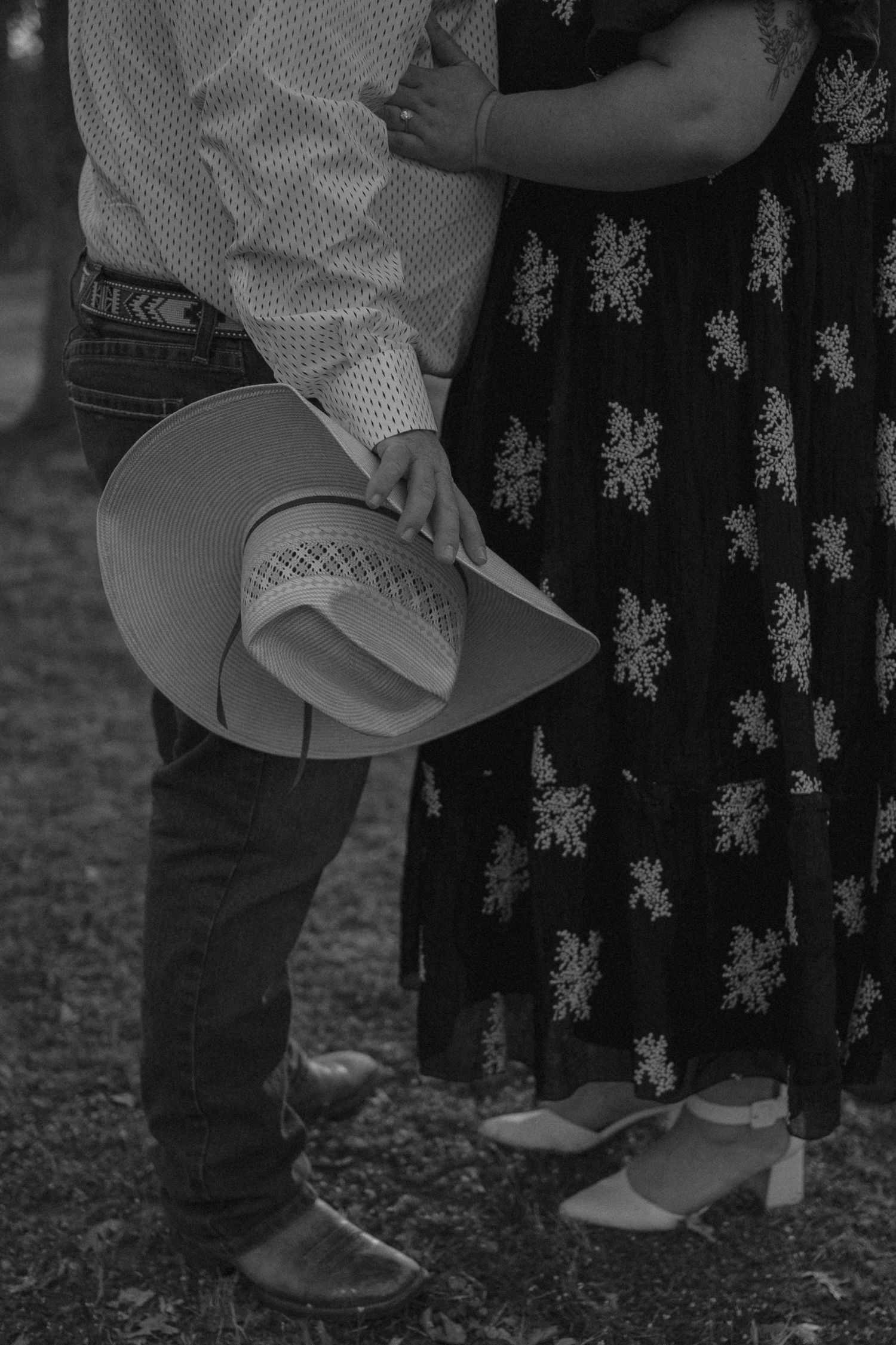 A black and white photo showing someone holding a cowboy hat while standing next to a person in a floral dress.