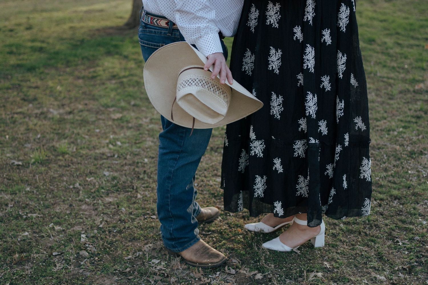 Close-up view of cowboy boots and western attire on two people standing on grass while holding a cowboy hat.