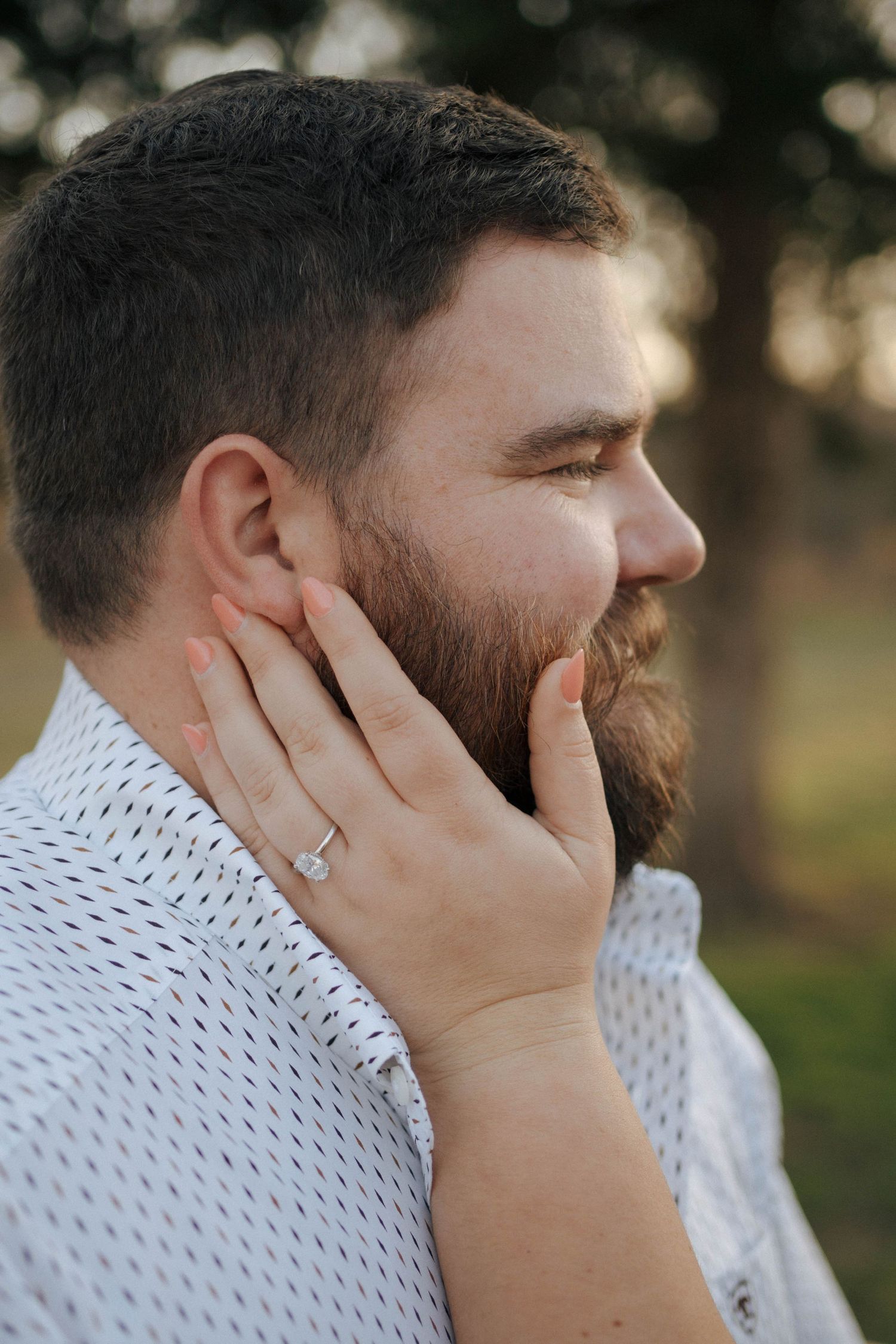 Hand resting on cheek with engagement ring showing in casual outdoor portrait showing a bearded profile.