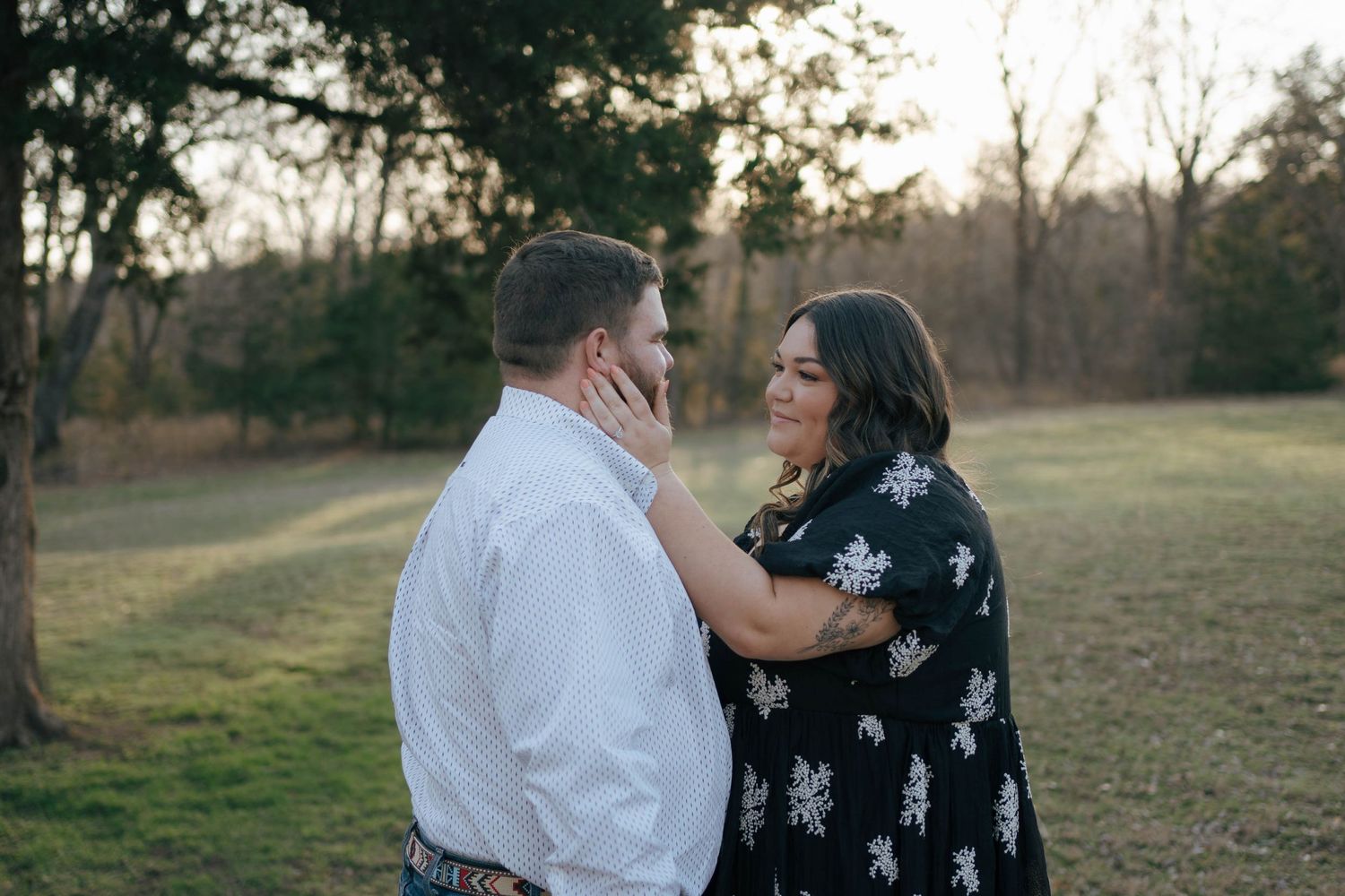 A loving couple embraces in a field during golden hour as sunlight filters through trees in the background.