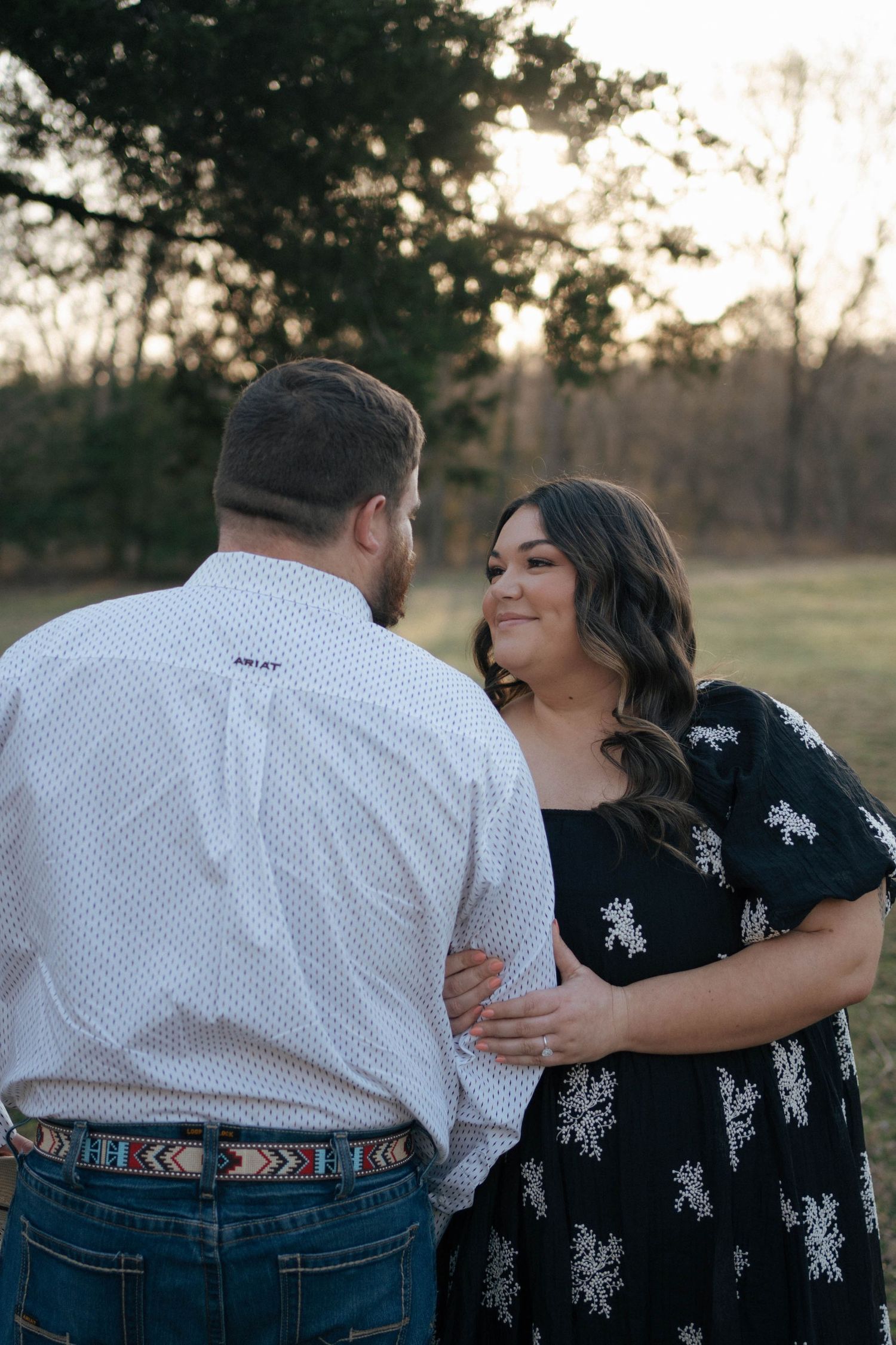 A couple embraces during golden hour outdoors with the sunset and trees visible in the background.