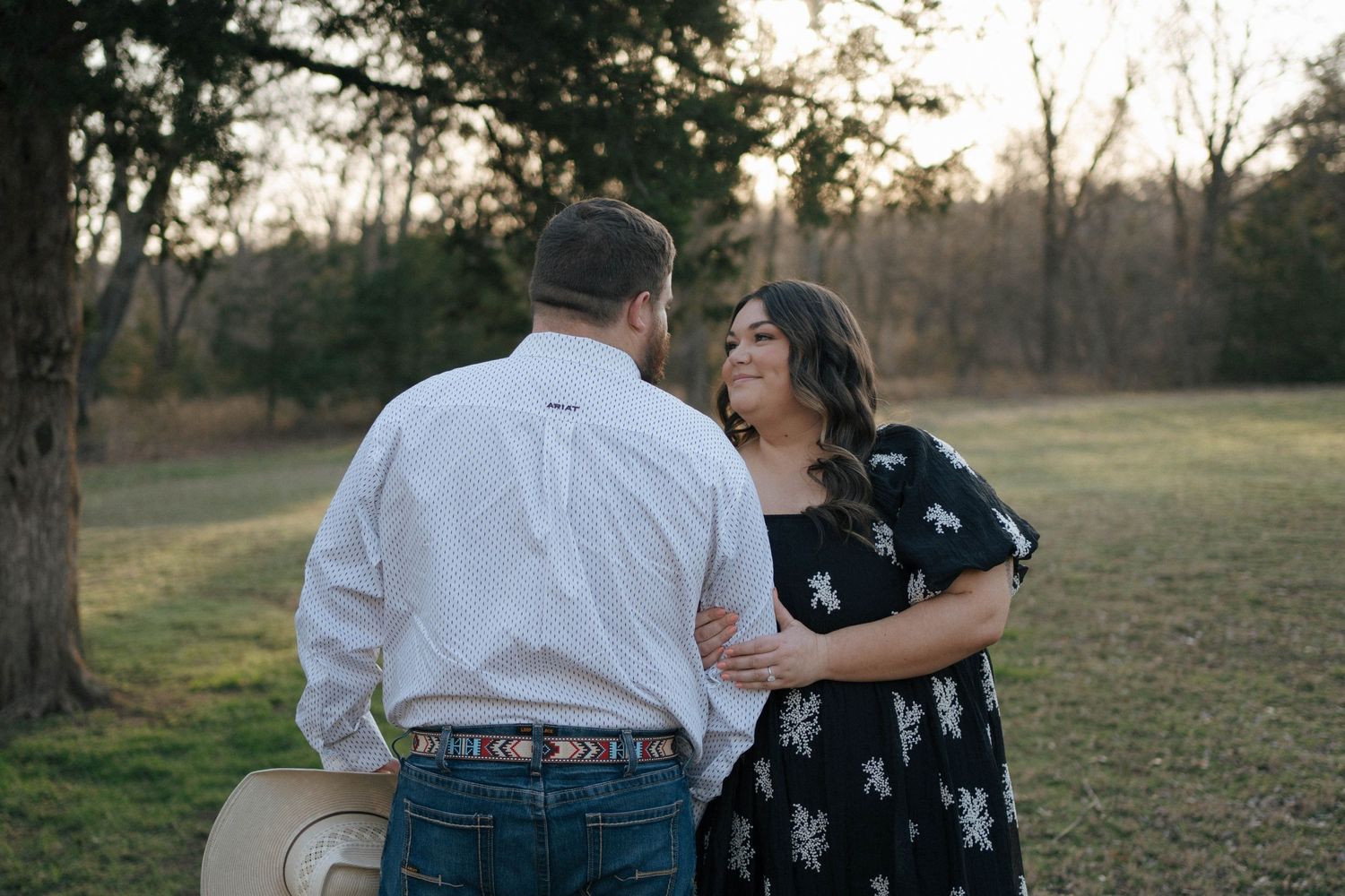 A couple in casual attire share a loving moment outdoors during sunset in a rustic field setting.