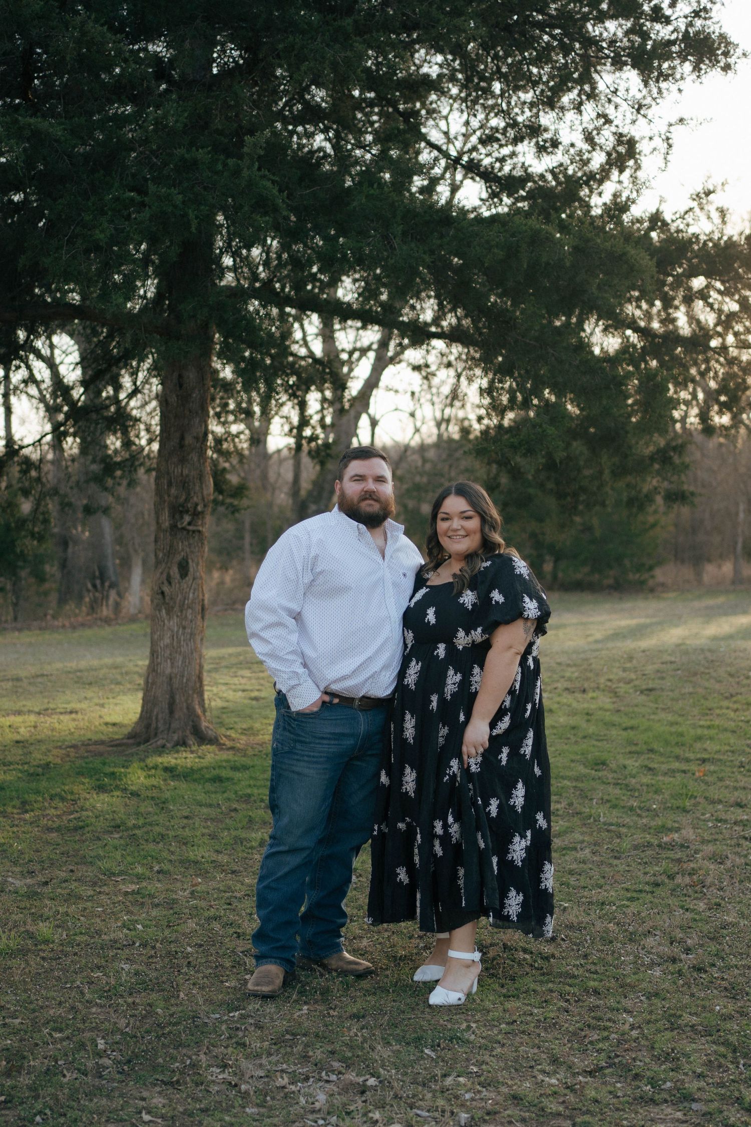 A couple poses together outdoors in a wooded area at sunset, wearing white and floral print outfits.
