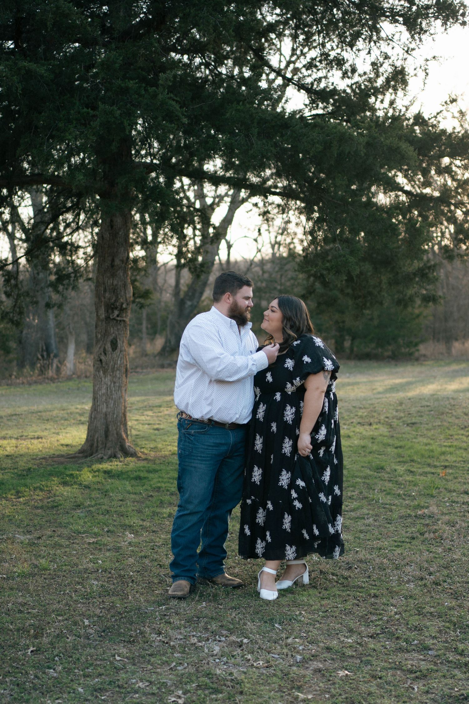 A couple shares a romantic moment outdoors at sunset beneath tall trees in a grassy field.