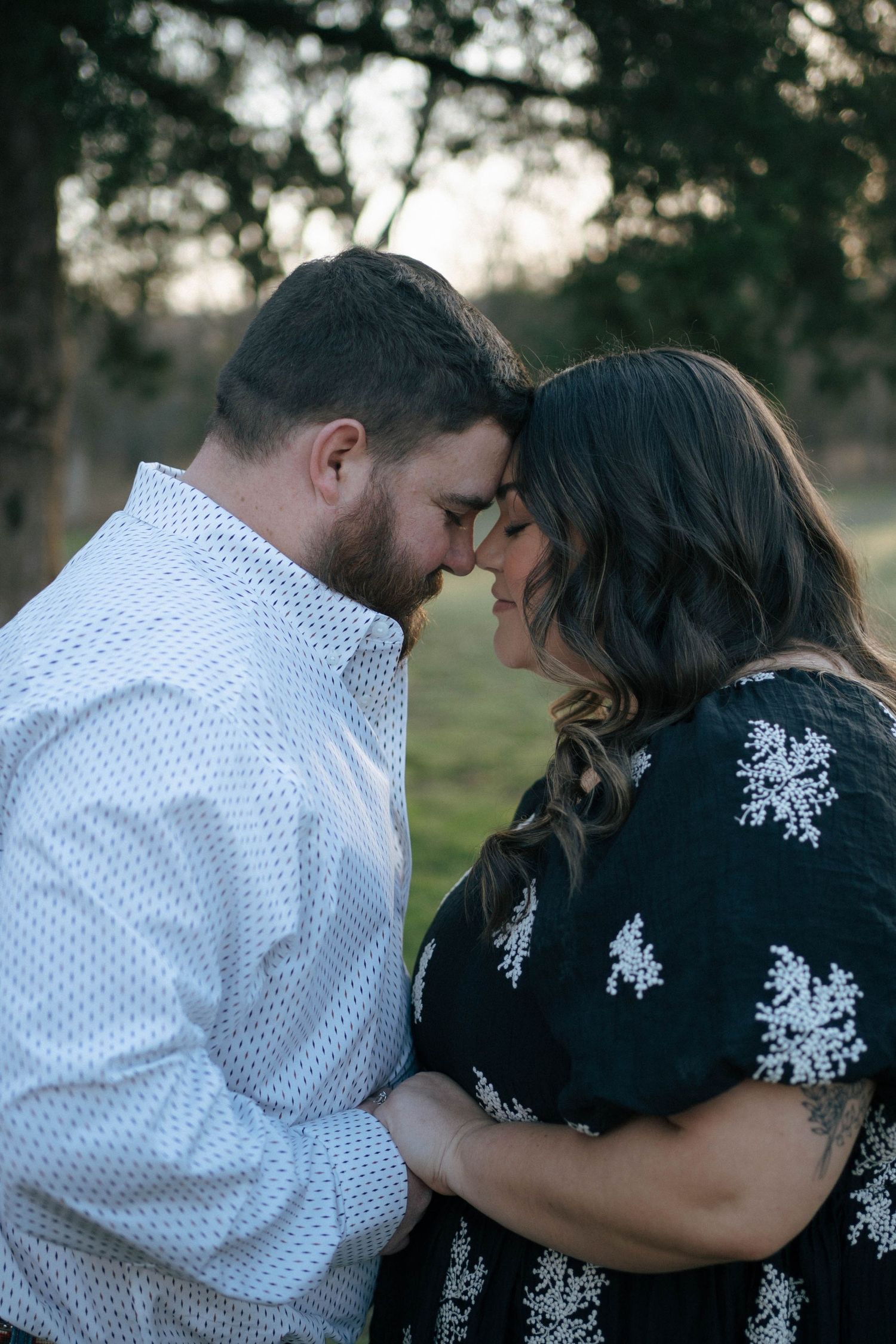 A romantic couple in a white polka dot shirt and black floral dress share an intimate moment at sunset outdoors.