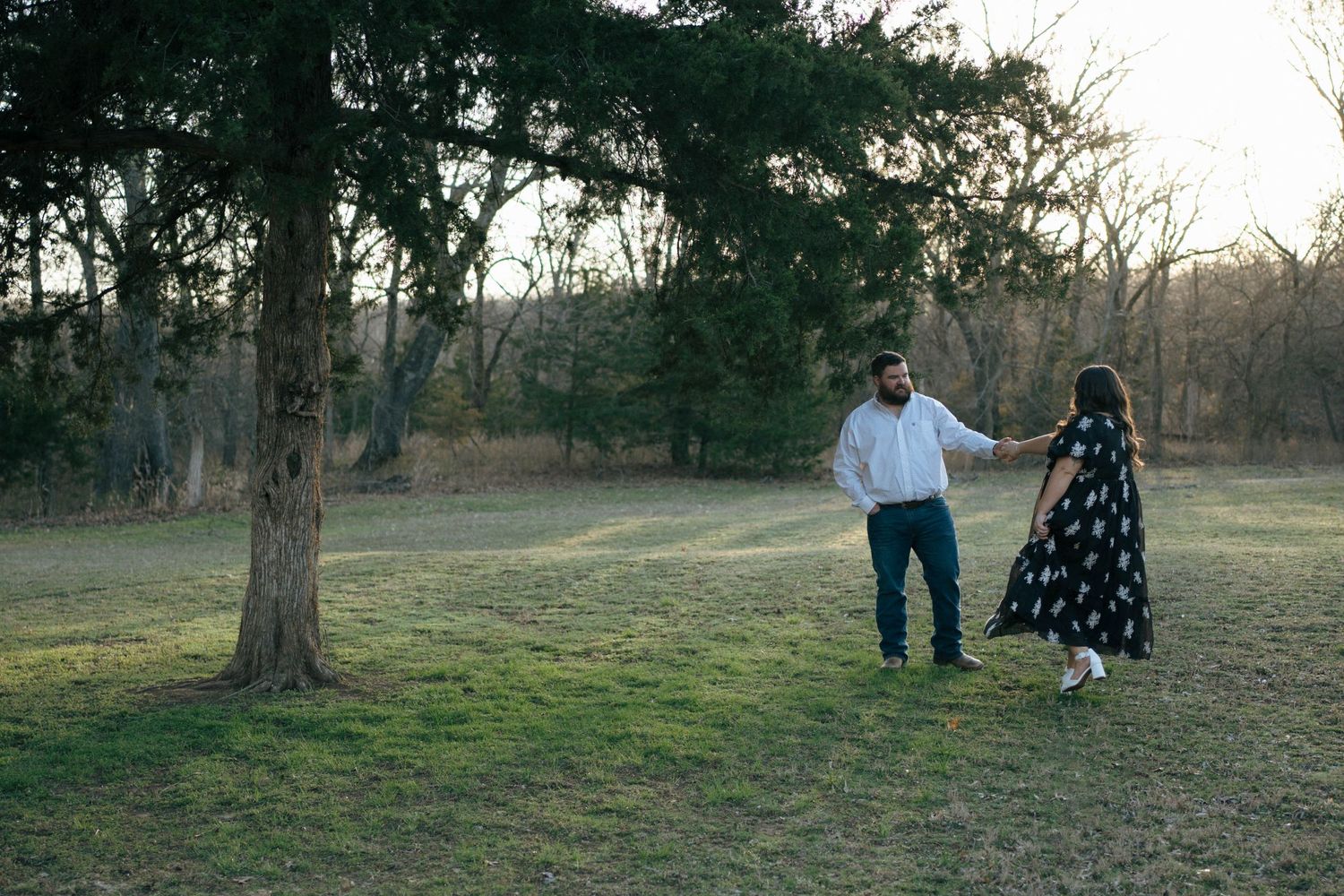 A couple stands together in a grassy field at sunset with trees in the background.