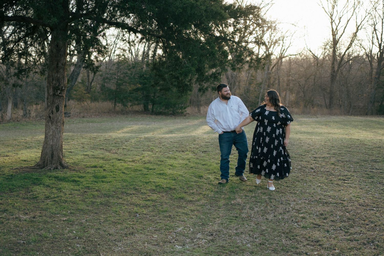 A couple walks hand in hand through a peaceful park setting during golden hour with bare trees in the background.