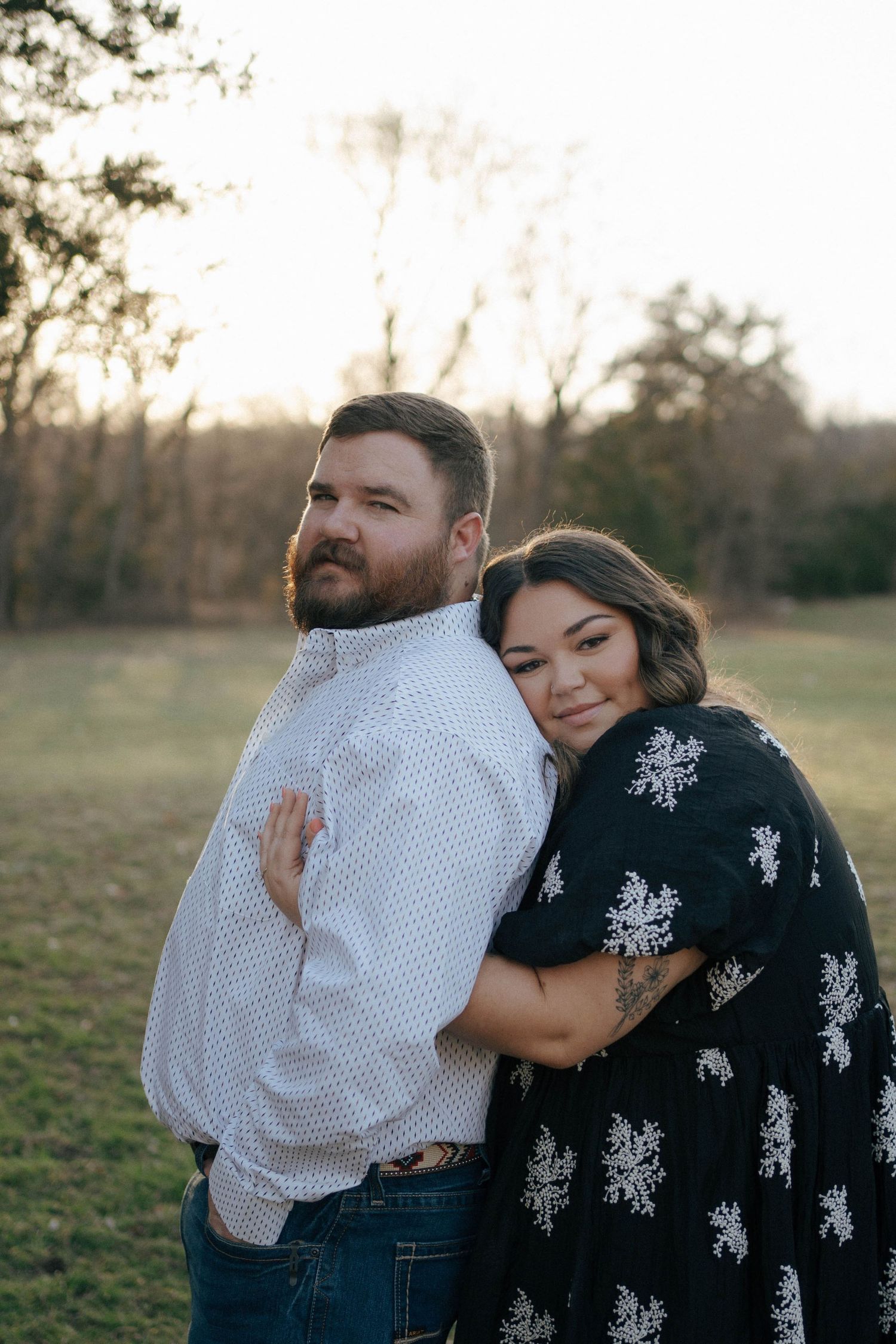 A couple embraces outdoors at sunset, with one wearing a white button-down shirt and the other in a black floral dress.