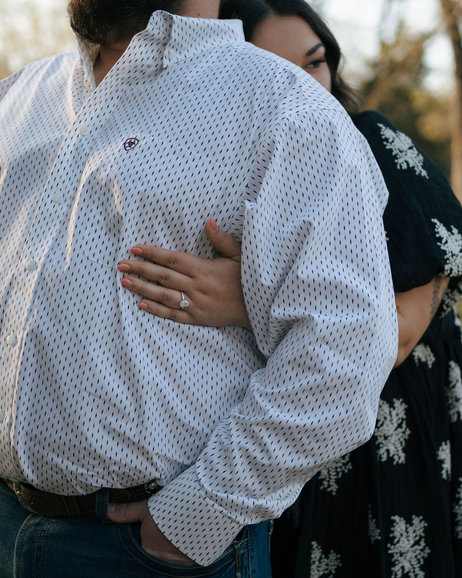 A close embrace shows hands resting on a white polka dot dress shirt against a dark floral dress in an outdoor setting.