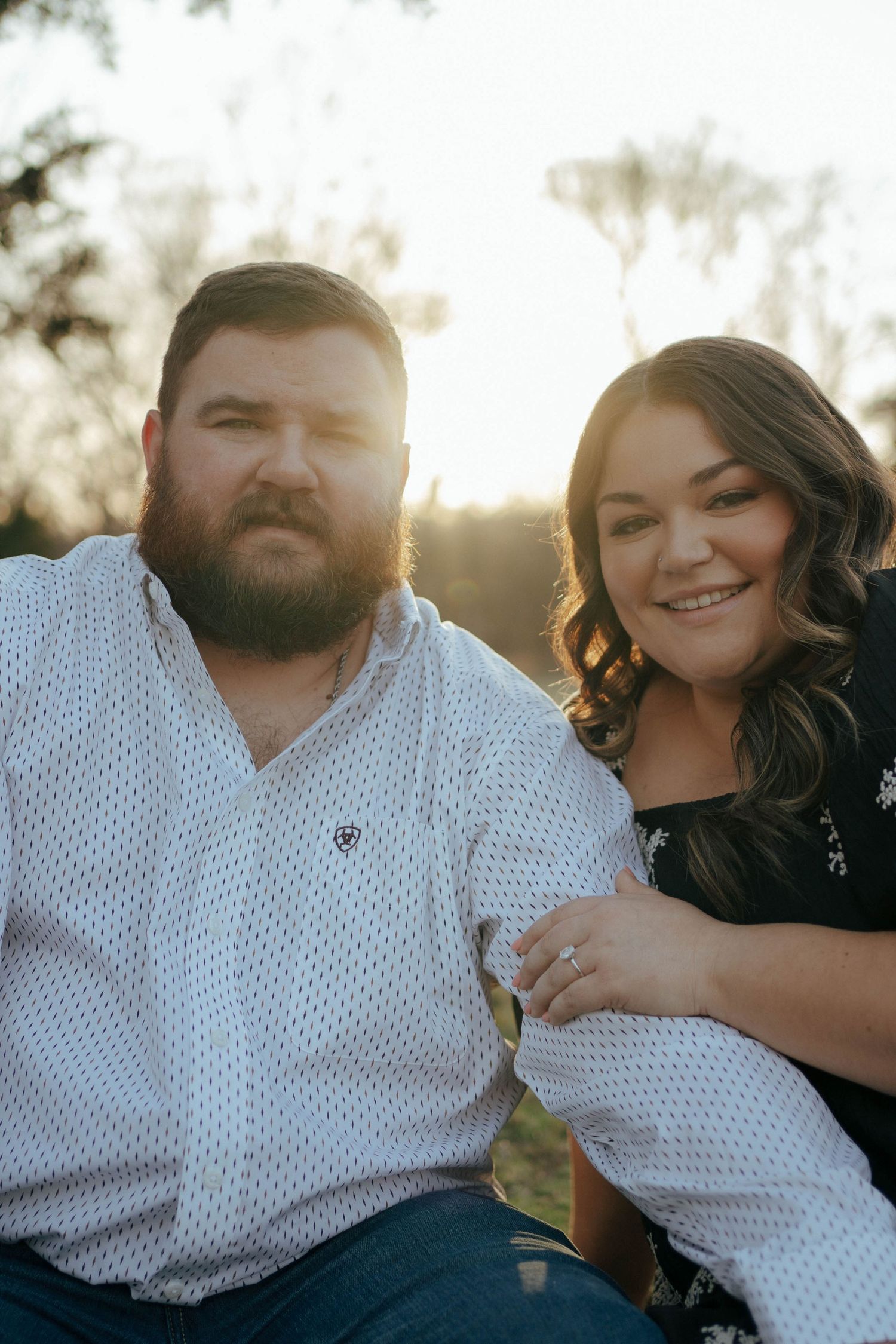 A couple poses together outdoors at sunset with sun flare in a casual portrait setting.