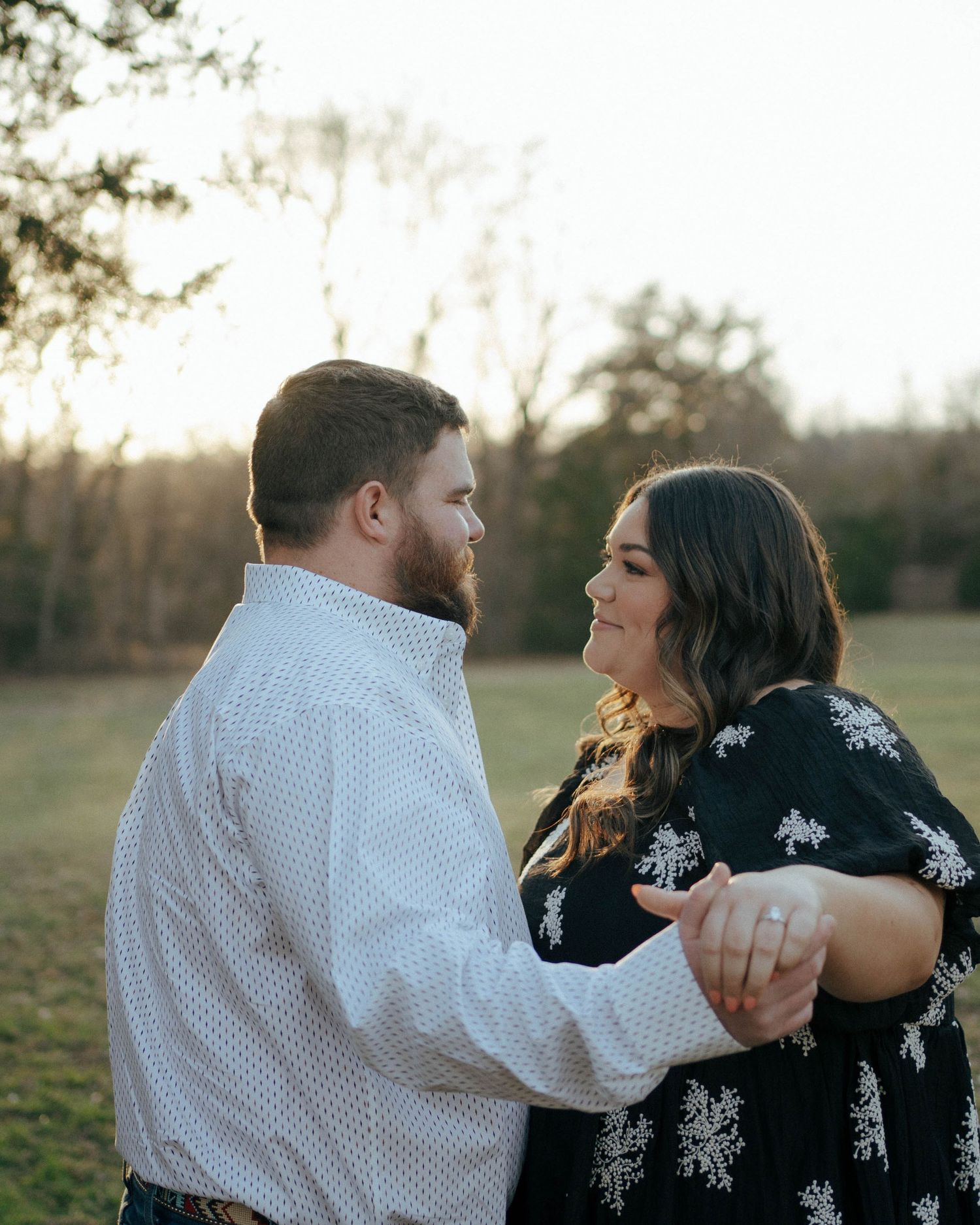 A couple shares a tender outdoor moment in a field at sunset while dressed in white polka dots and black floral prints.