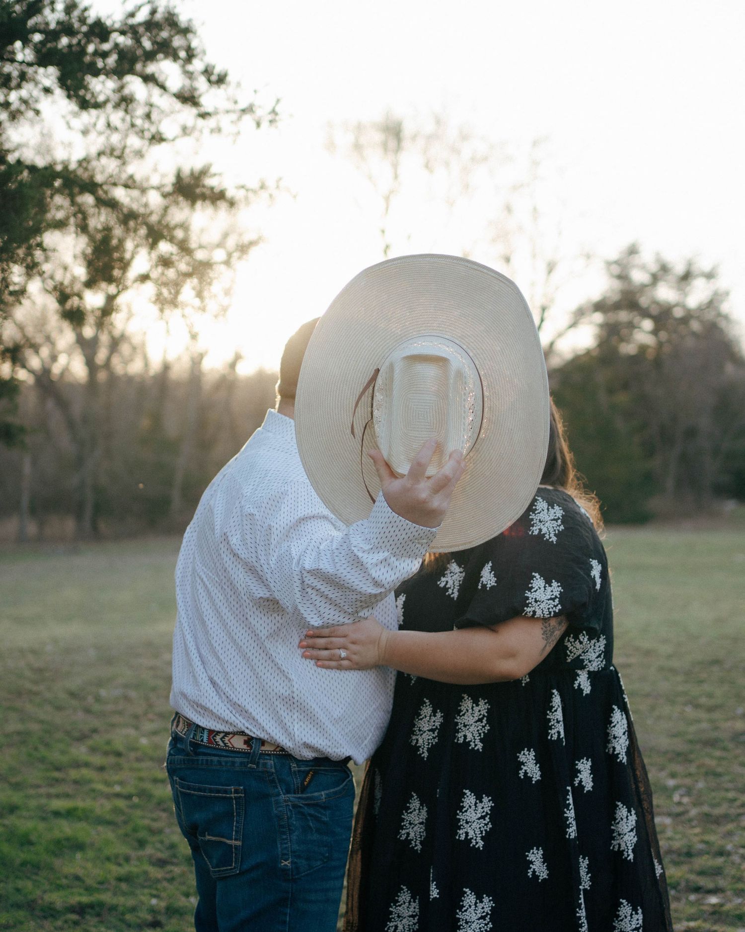 Couple sharing an intimate moment at sunset in a field while one wears a large sun hat and floral dress.