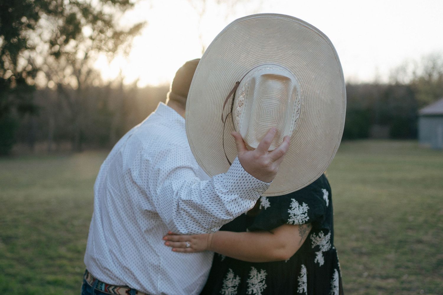A romantic moment in a field at sunset with a couple sharing an intimate embrace behind a wide-brimmed hat.