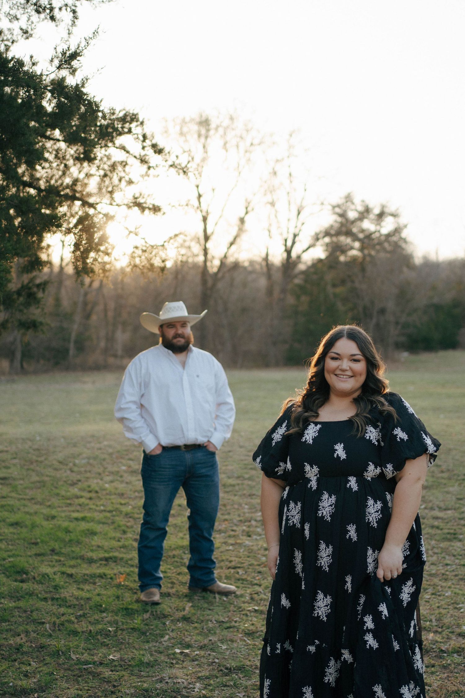 A couple poses in a rustic outdoor setting at sunset with a man in western attire and woman in a floral dress.