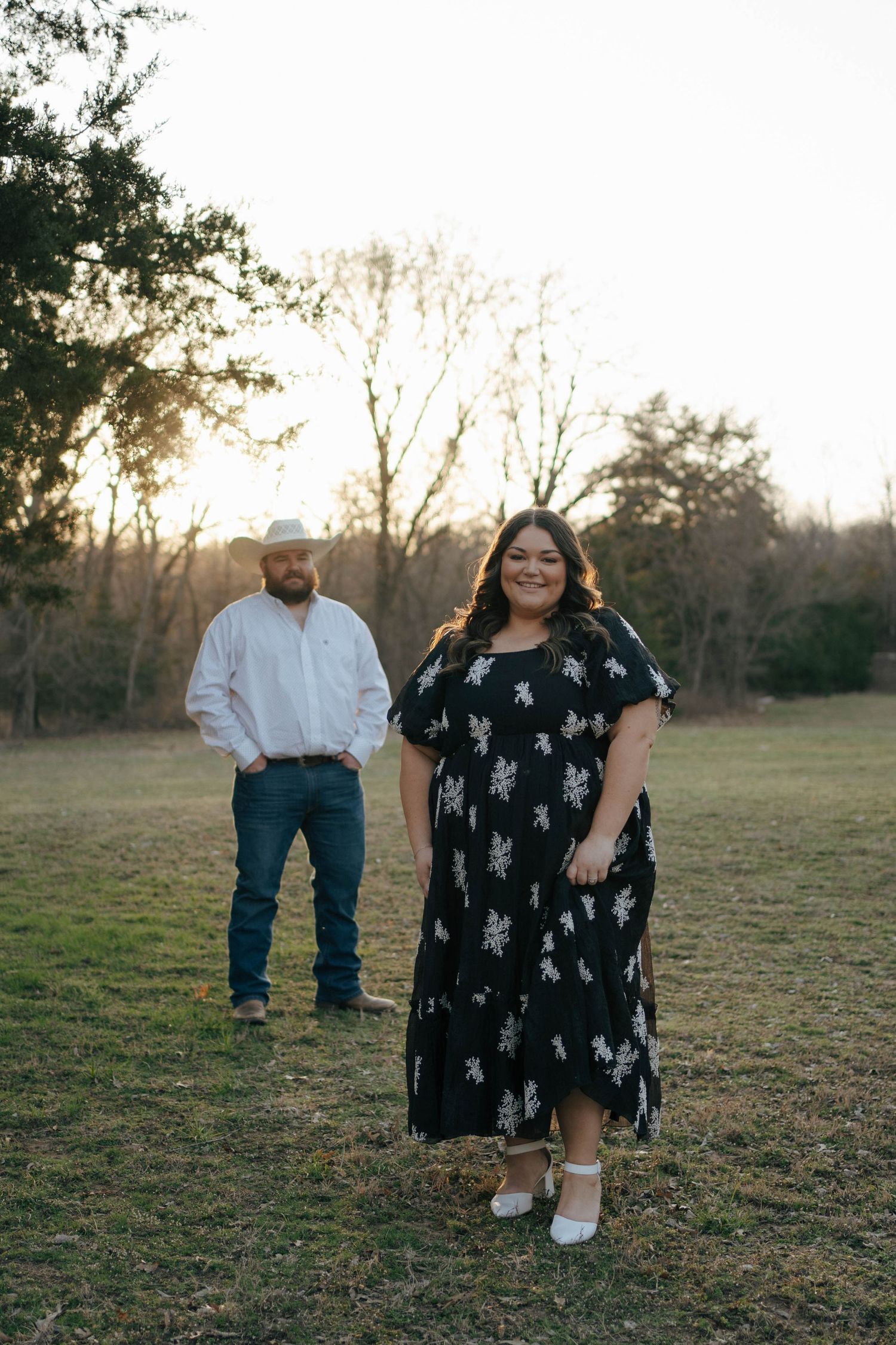 A couple stands in a grassy field at sunset wearing western-inspired outfits with casual country styling.
