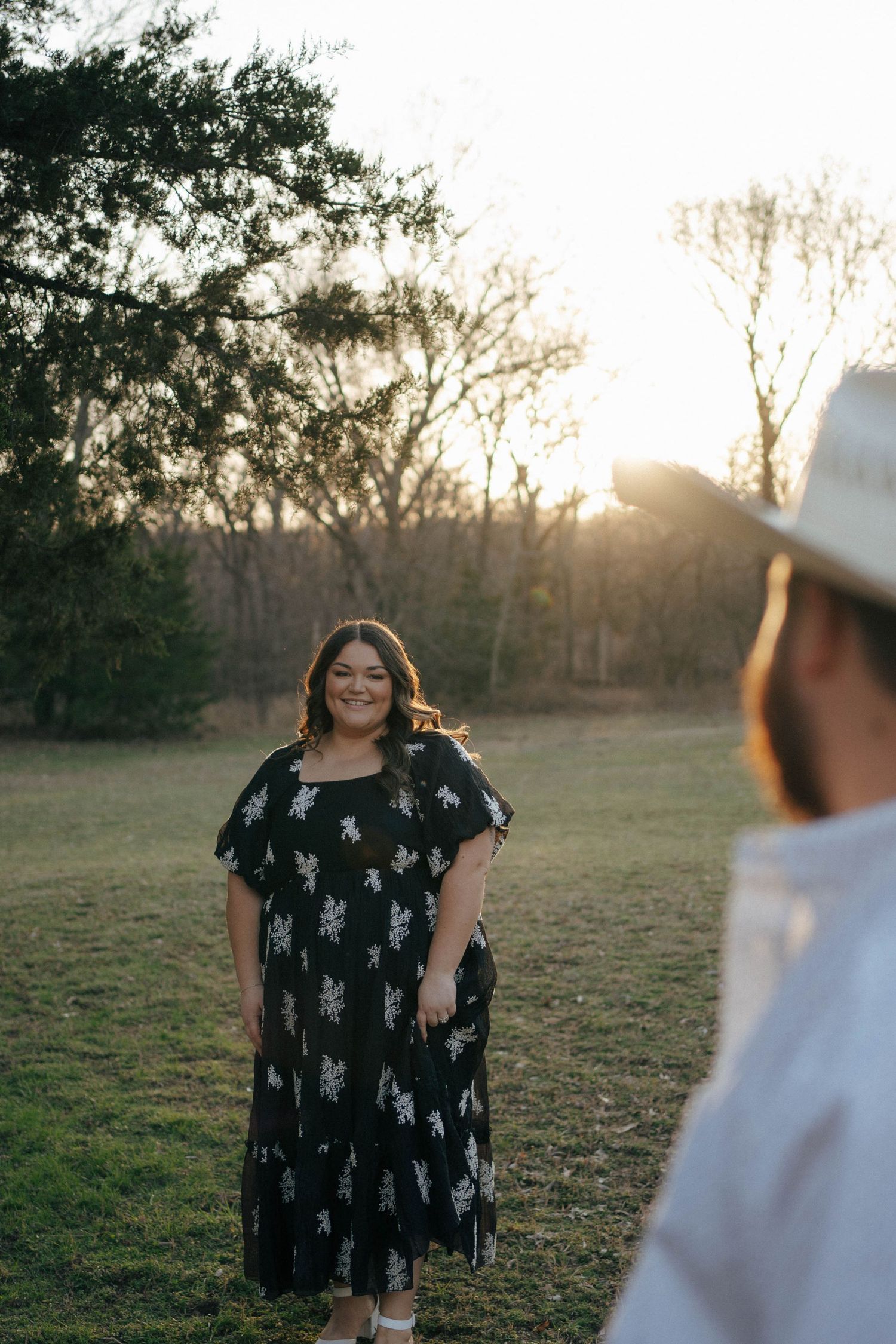 A person in a black floral dress stands in a field at sunset while someone in white looks on from the foreground.