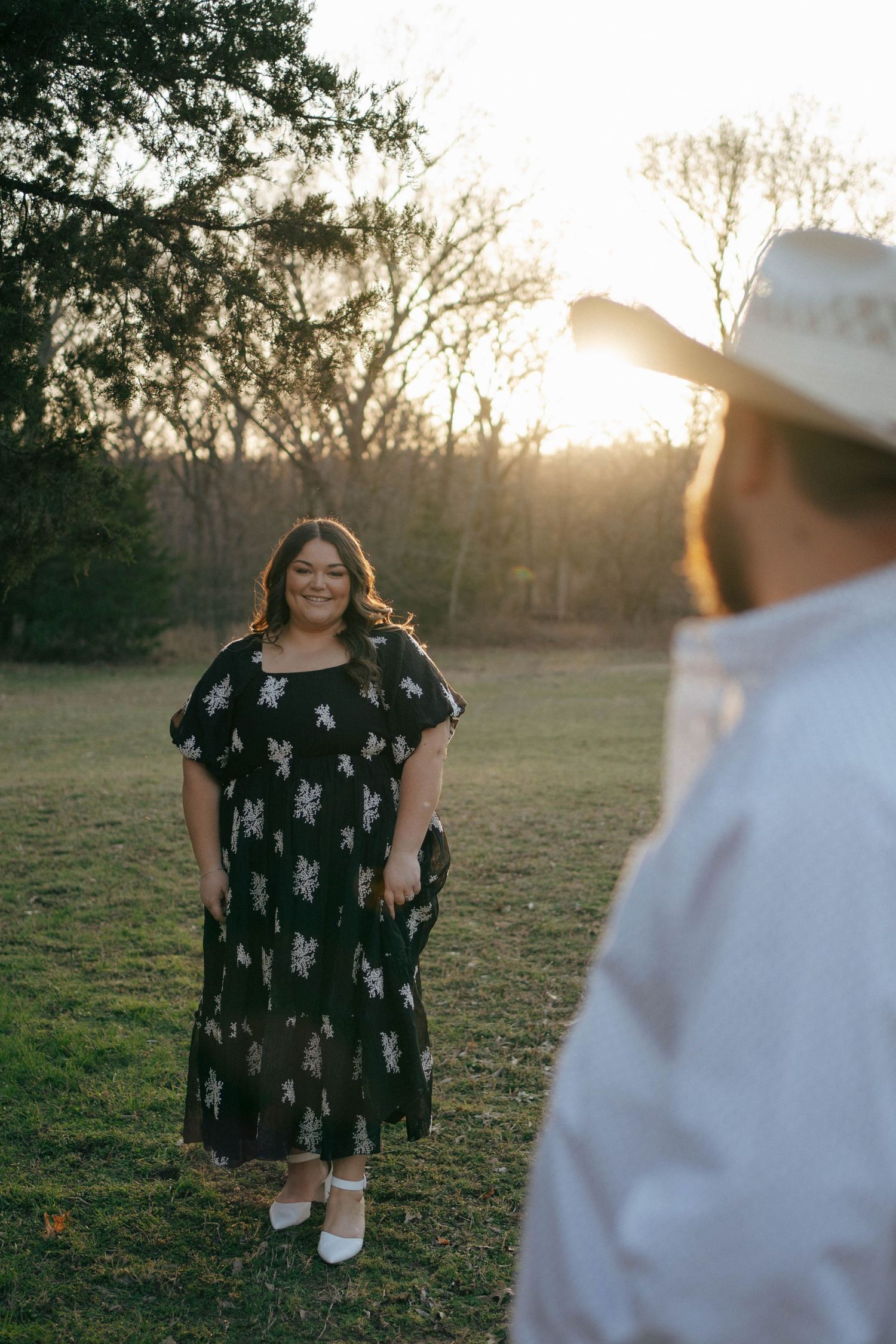 A person wearing a black patterned dress poses outdoors at golden hour while being photographed.