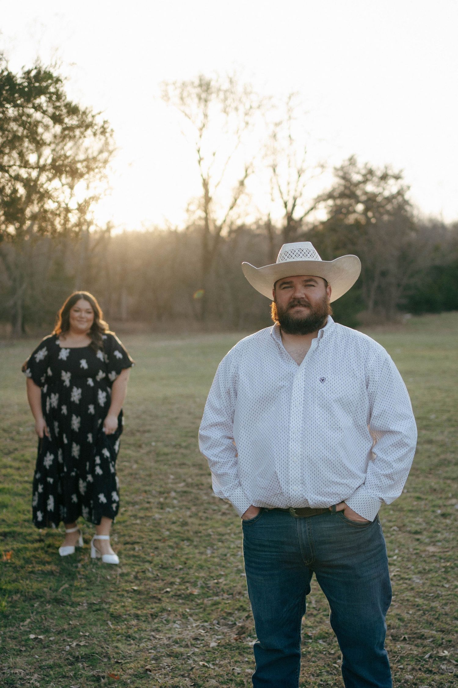 A couple poses in a field at sunset with a person in a polka dot dress in the background and a cowboy up front.