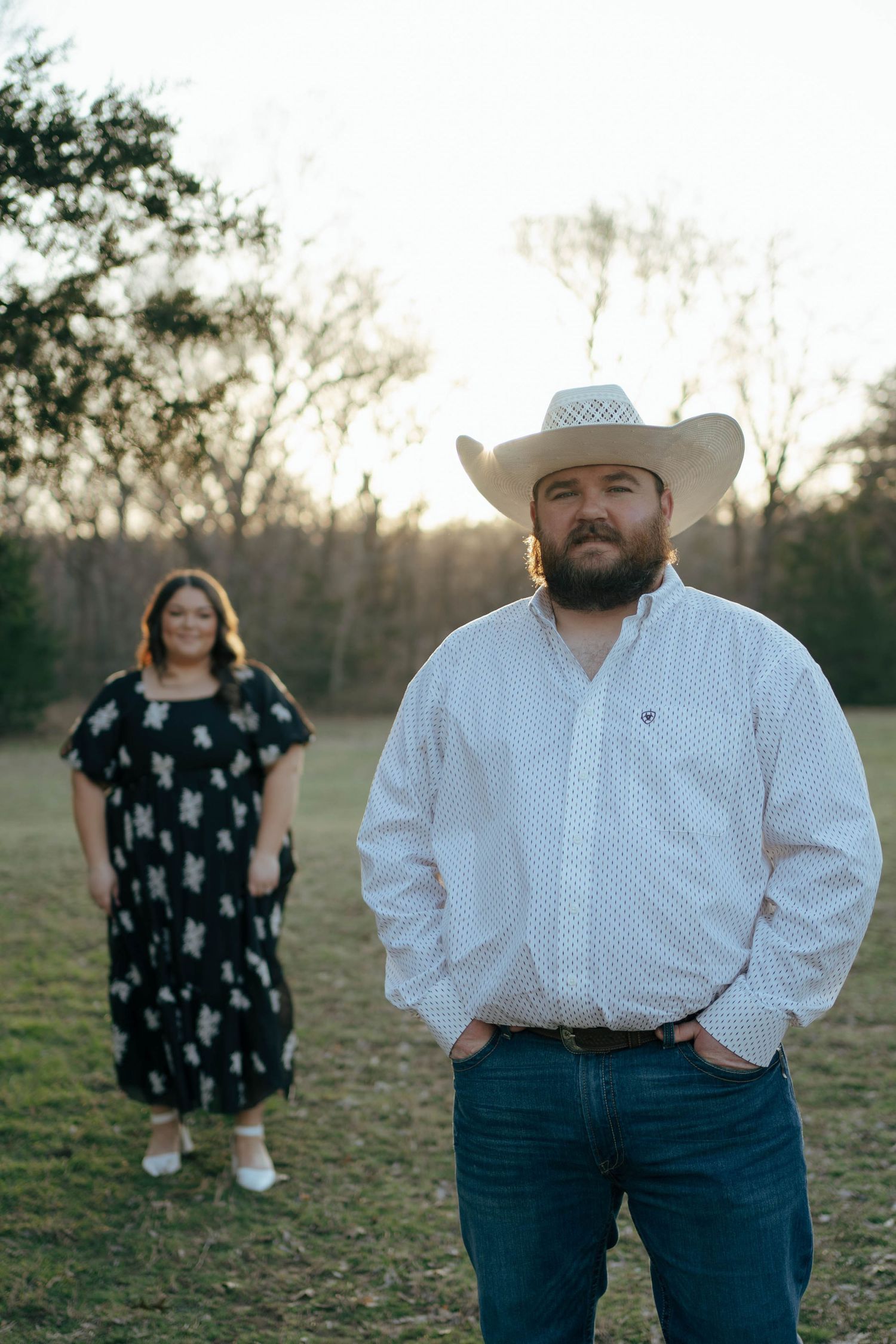 A country-western couple poses in an open field at dusk with the person in white wearing a cowboy hat.
