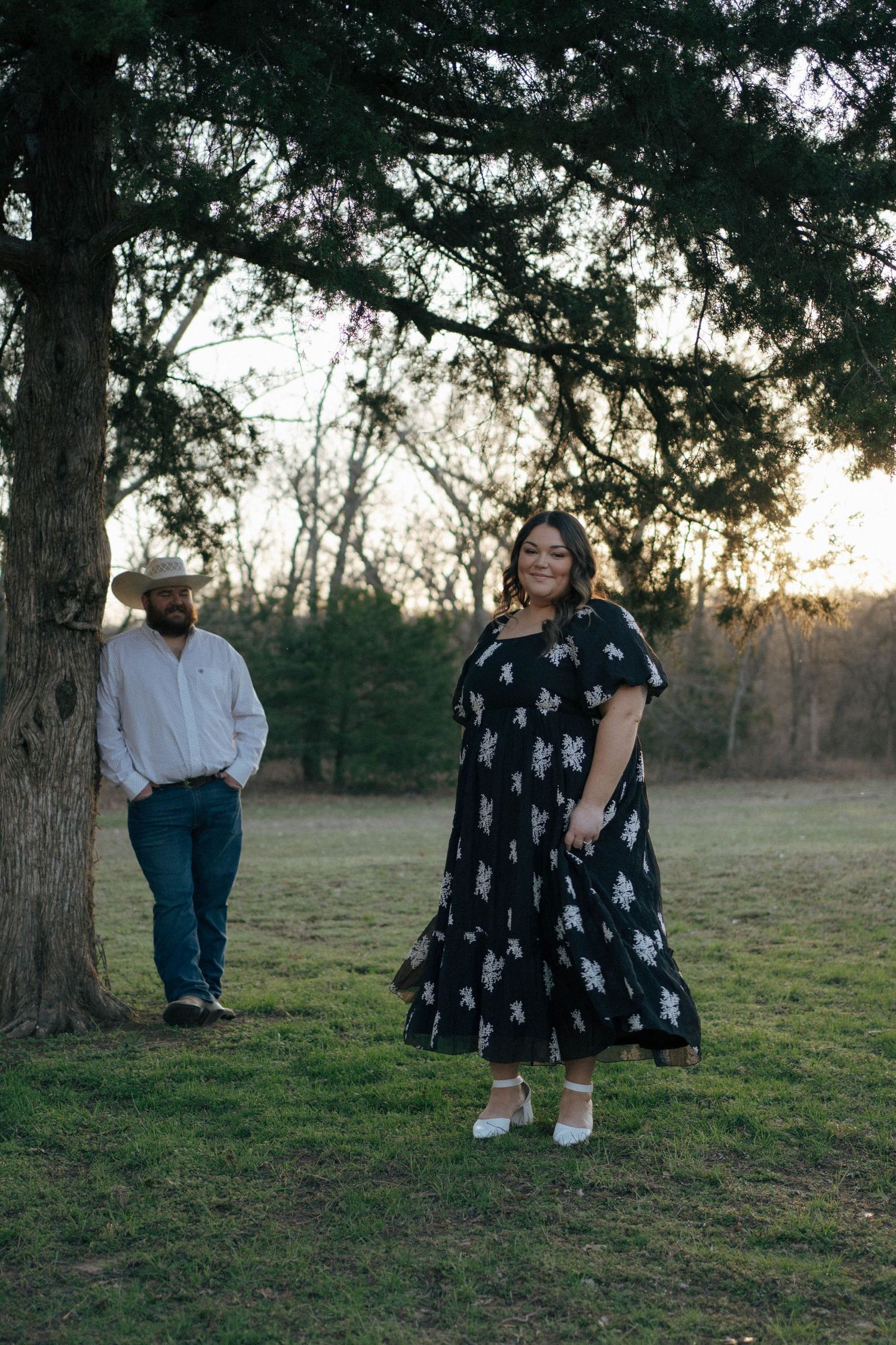A couple stands beneath tall trees at dusk with a man in cowboy attire and woman in a patterned maxi dress.