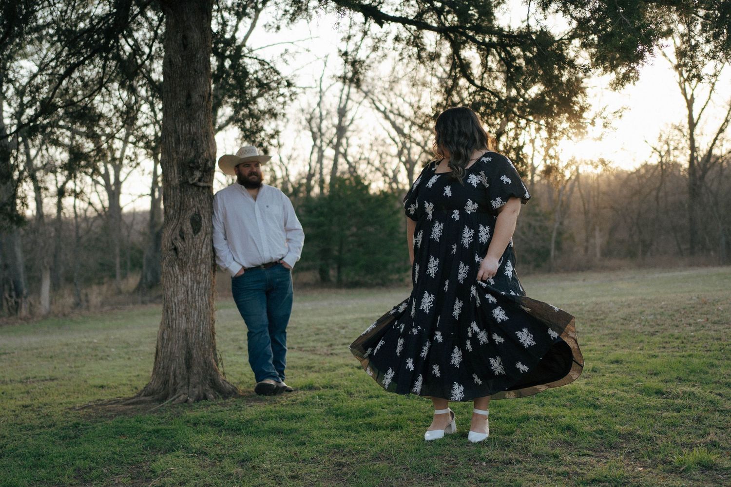 Two people in a park at sunset with one wearing a white shirt and the other in a twirling polka dot dress.