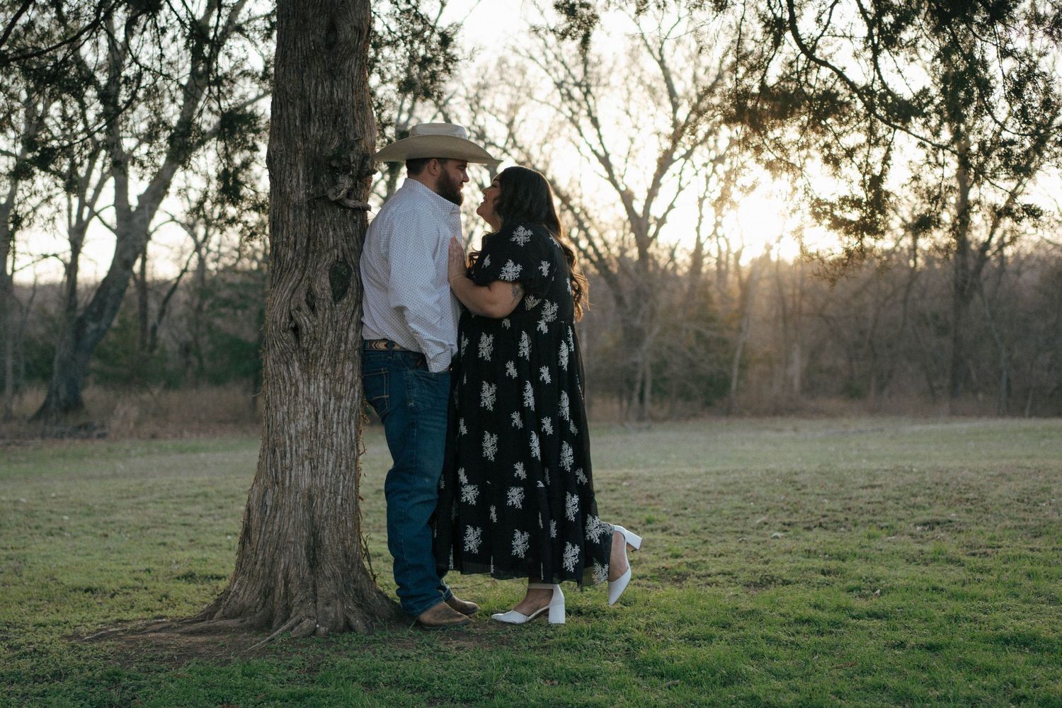 A couple stands together under a tree at sunset in a serene outdoor setting.