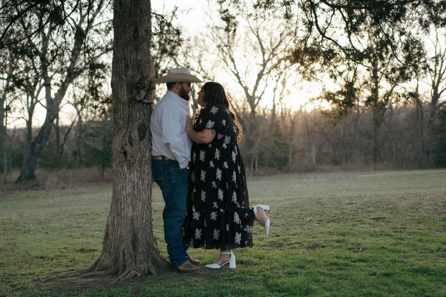 Three people standing by a tree in a grassy field at sunset on a tranquil evening.