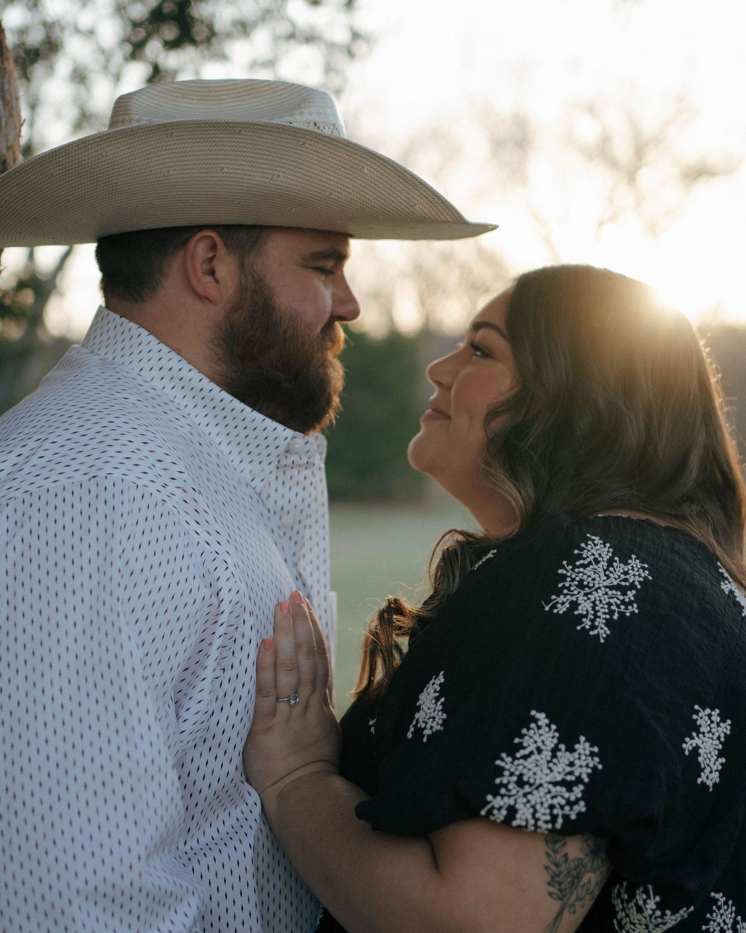 A couple shares an intimate moment at sunset, with one wearing a cowboy hat and white shirt and the other in a floral dress.