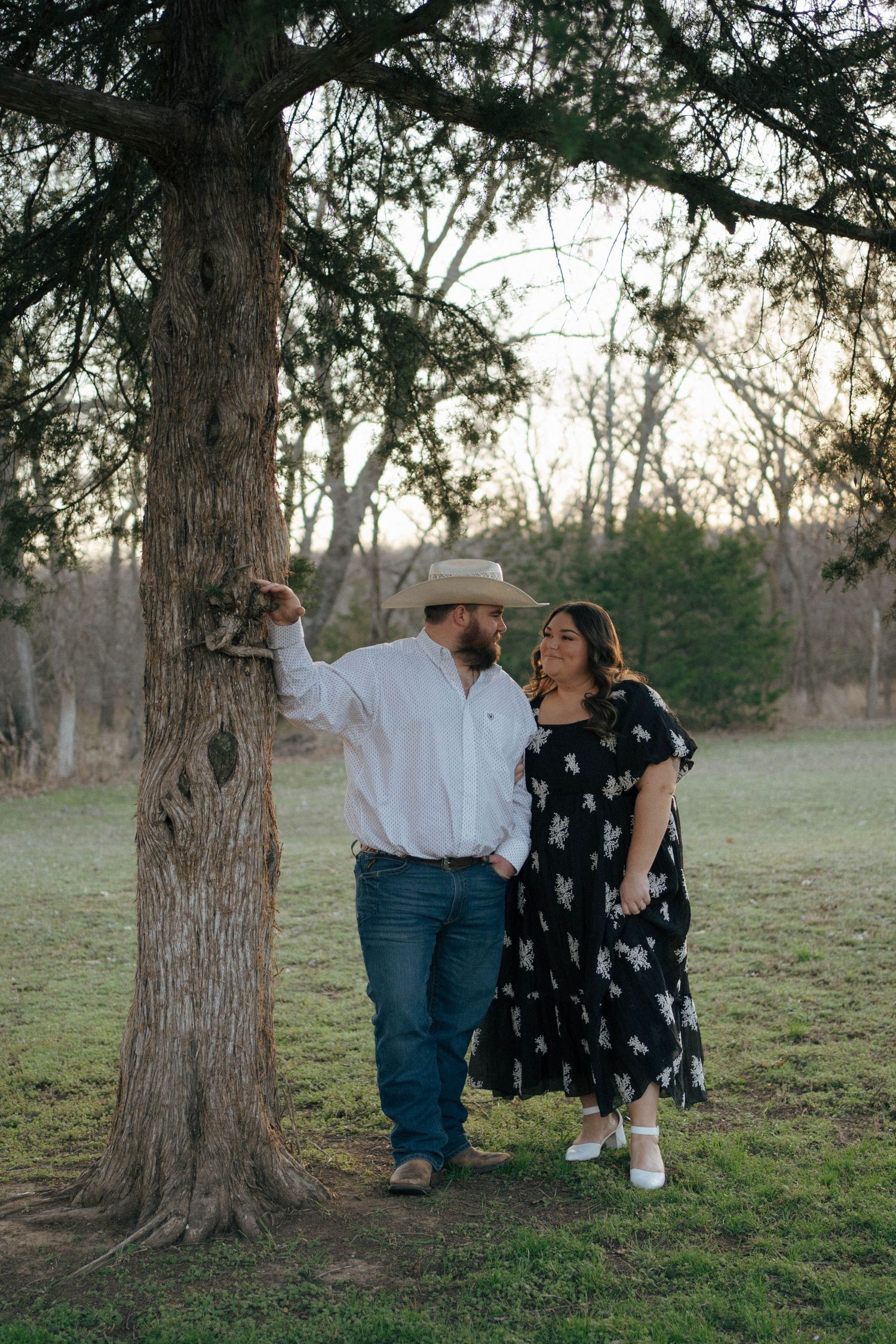 A couple stands together under a large tree at sunset in a rustic outdoor setting.