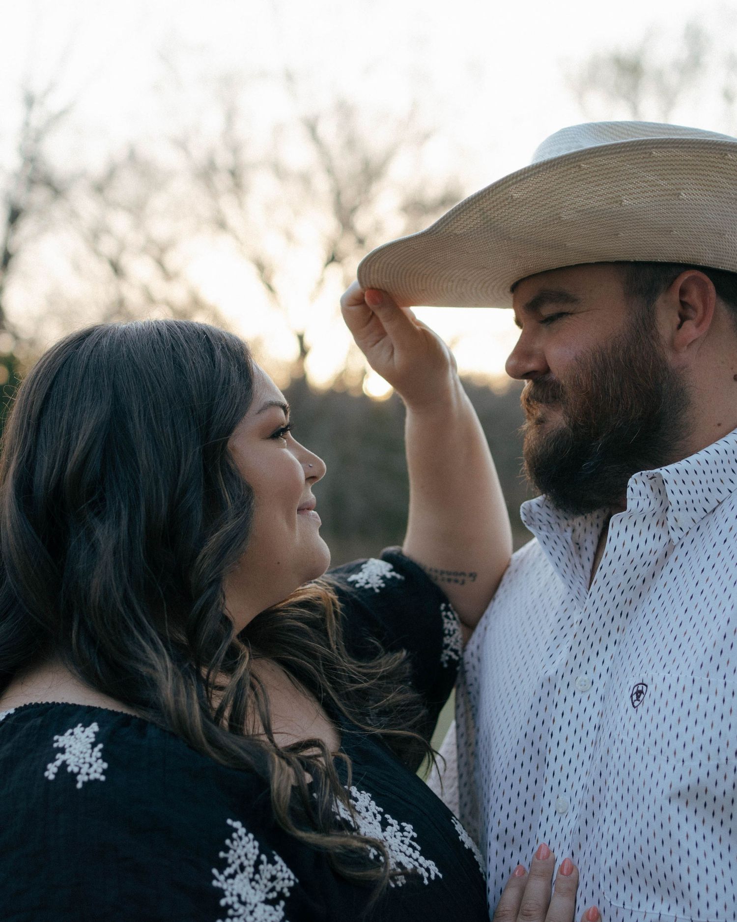 A romantic couple shares a tender moment at sunset, with the person wearing a cowboy hat and white shirt.