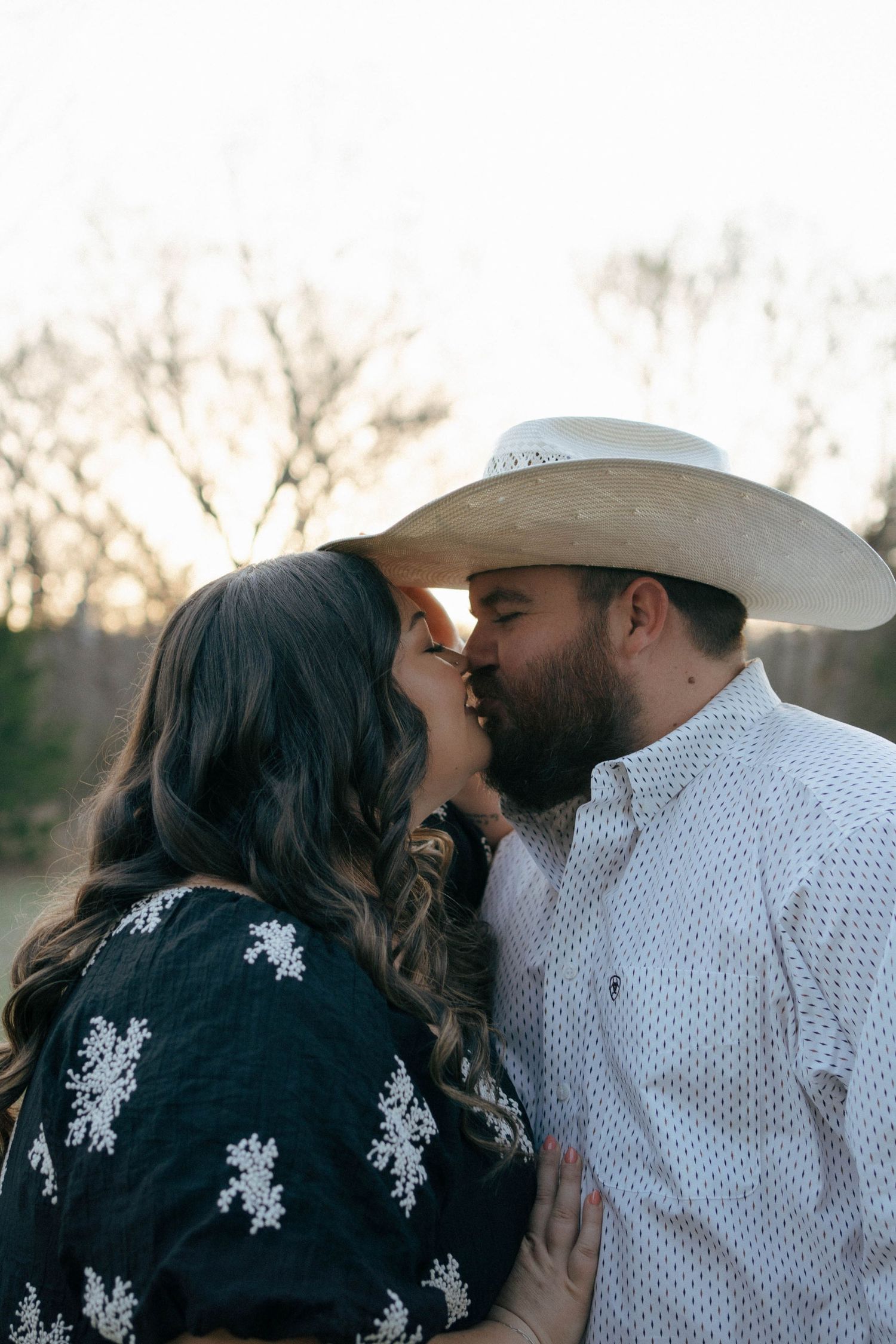 A romantic couple shares a tender kiss at sunset, with one wearing a cowboy hat and white shirt and the other in a dark patterned top.