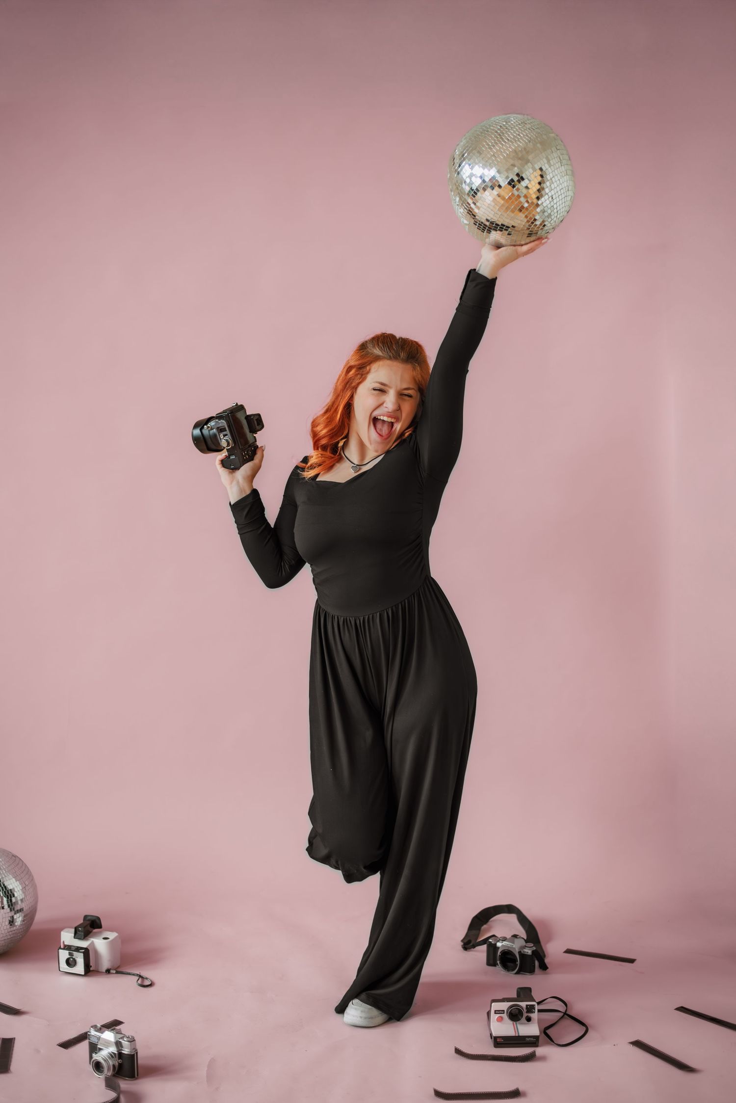 A joyful photographer jumps with camera and disco ball against pink studio backdrop.