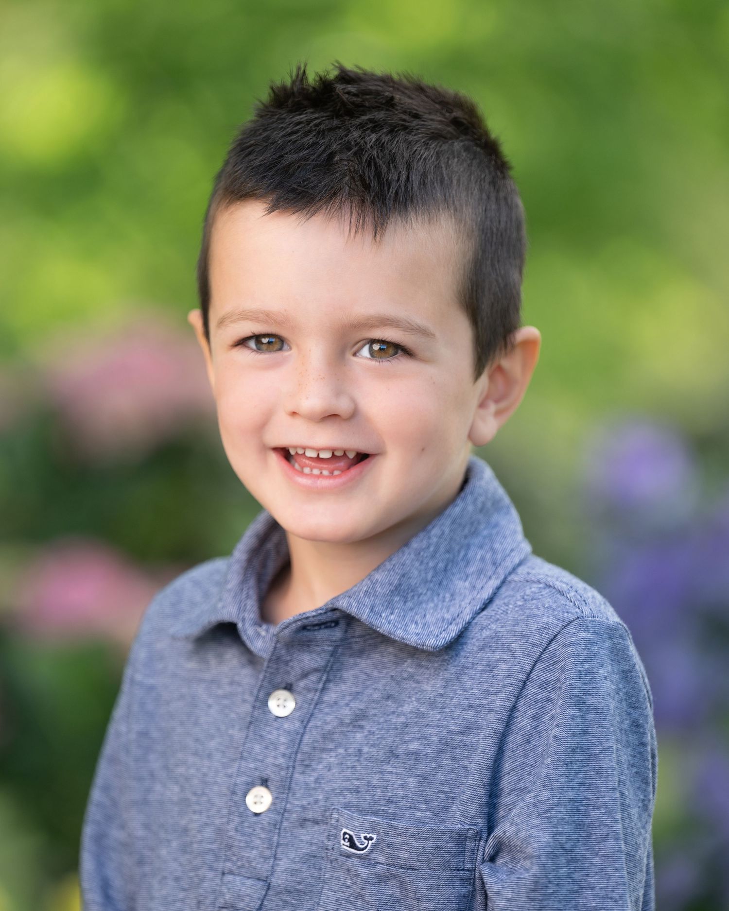 Young student wearing navy cardigan and light colored button-up shirt smiles while posing outdoors against blurred garden background.