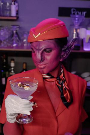 Pink alien dressed like a flight attendant making drinks.