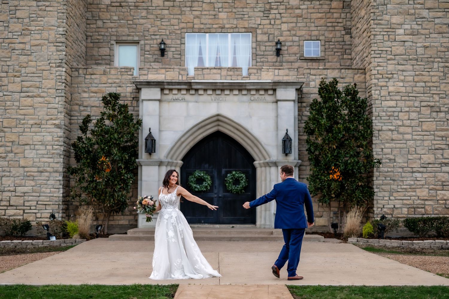 Two people in formal attire walk along a stone path leading to a gothic-style church entrance with arched doorway.