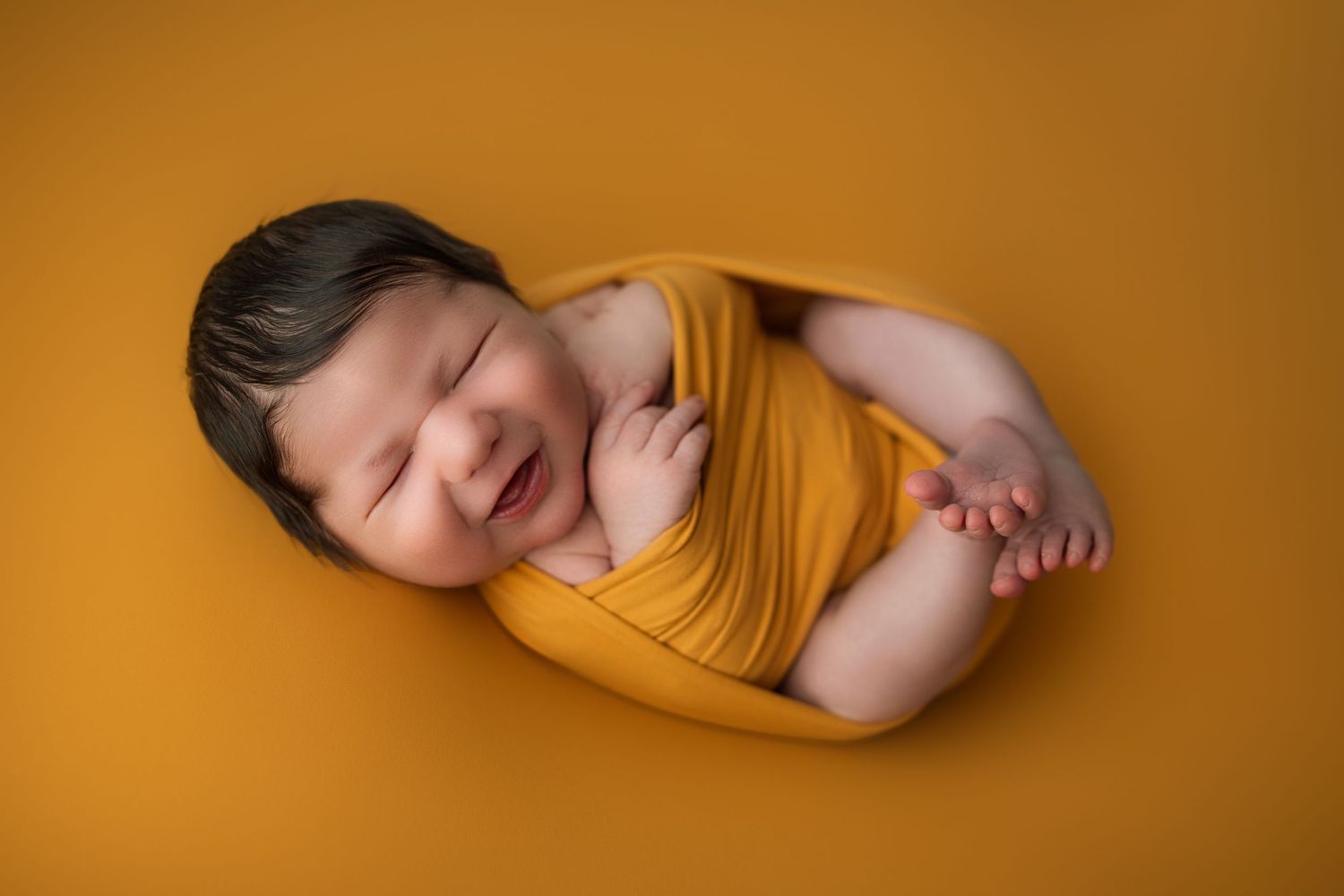 Professional newborn photography session with soft wraps against a warm mustard yellow background.