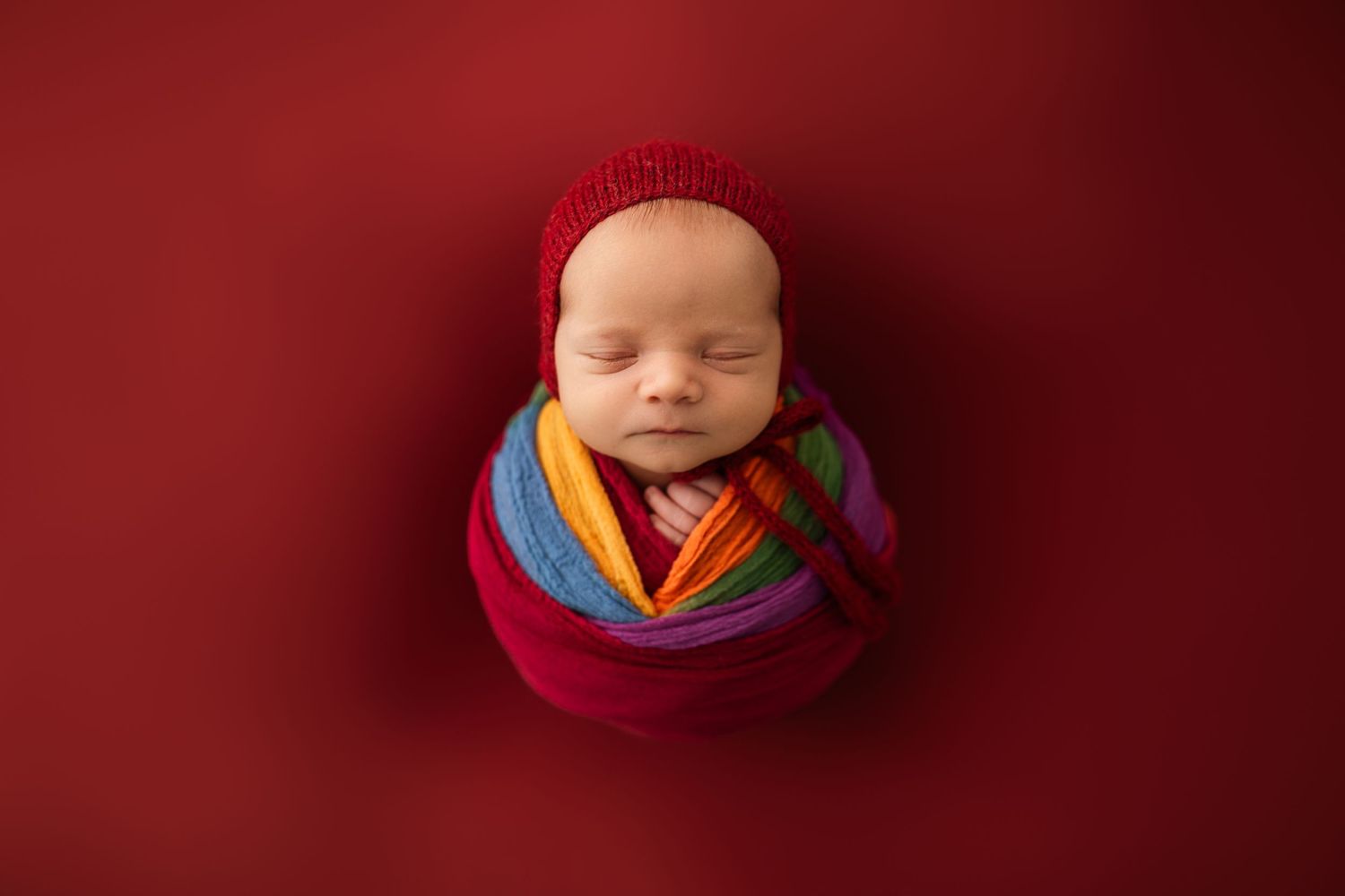 Creative newborn photography with vibrant red backdrop and colorful knit accessories.
