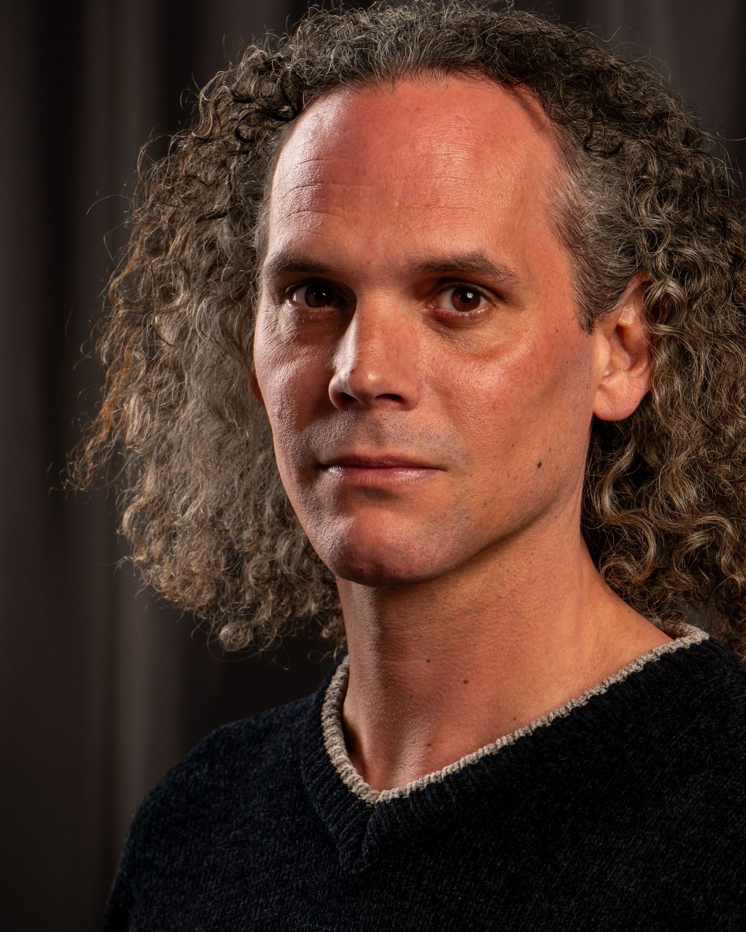 A person with curly shoulder-length hair wearing a black sweater poses against a dark background in a studio portrait.