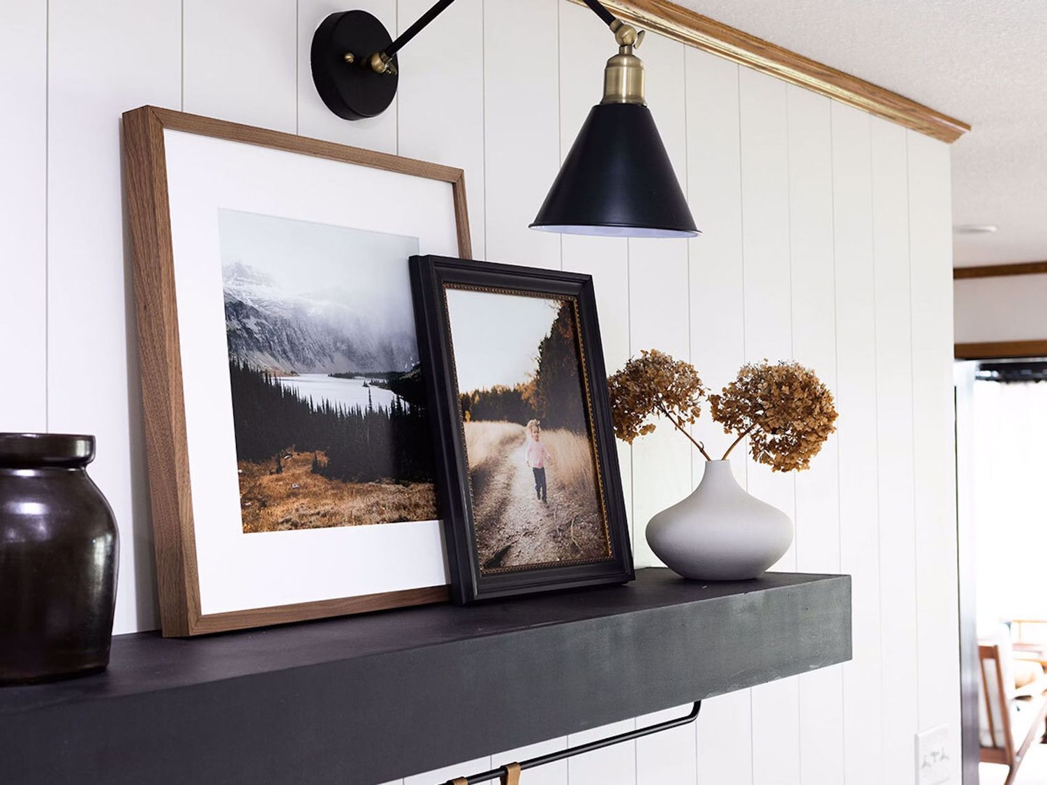 A black floating shelf displays framed landscape photographs and a vase with dried flowers, accented by a black wall sconce.