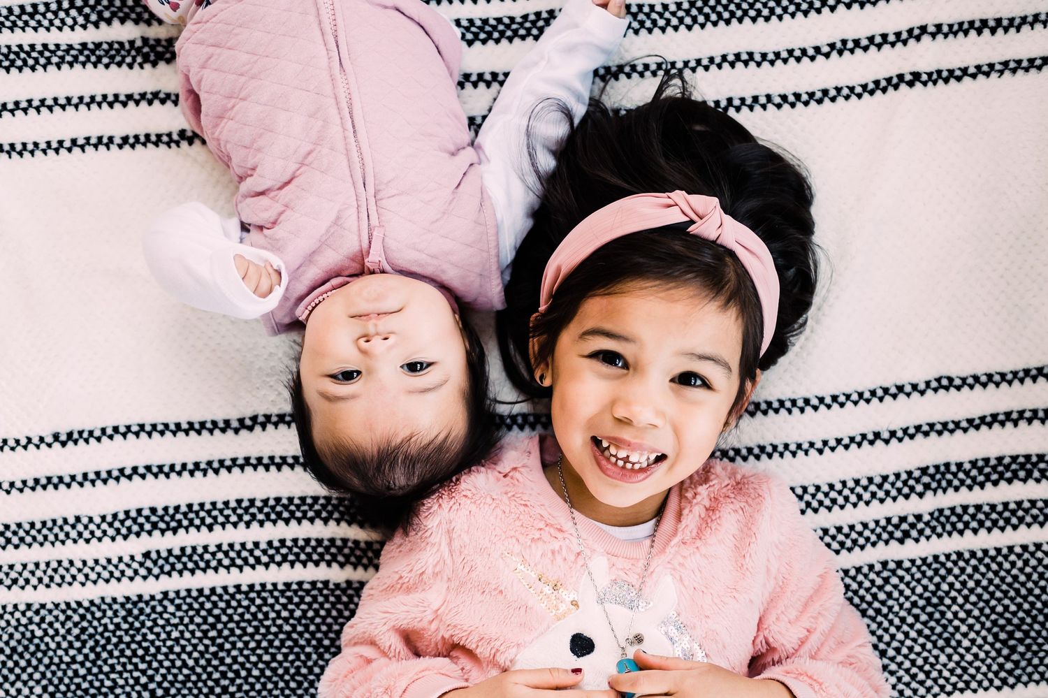 Two children lying on a striped blanket wearing pink sweaters and smiling while looking up at camera.