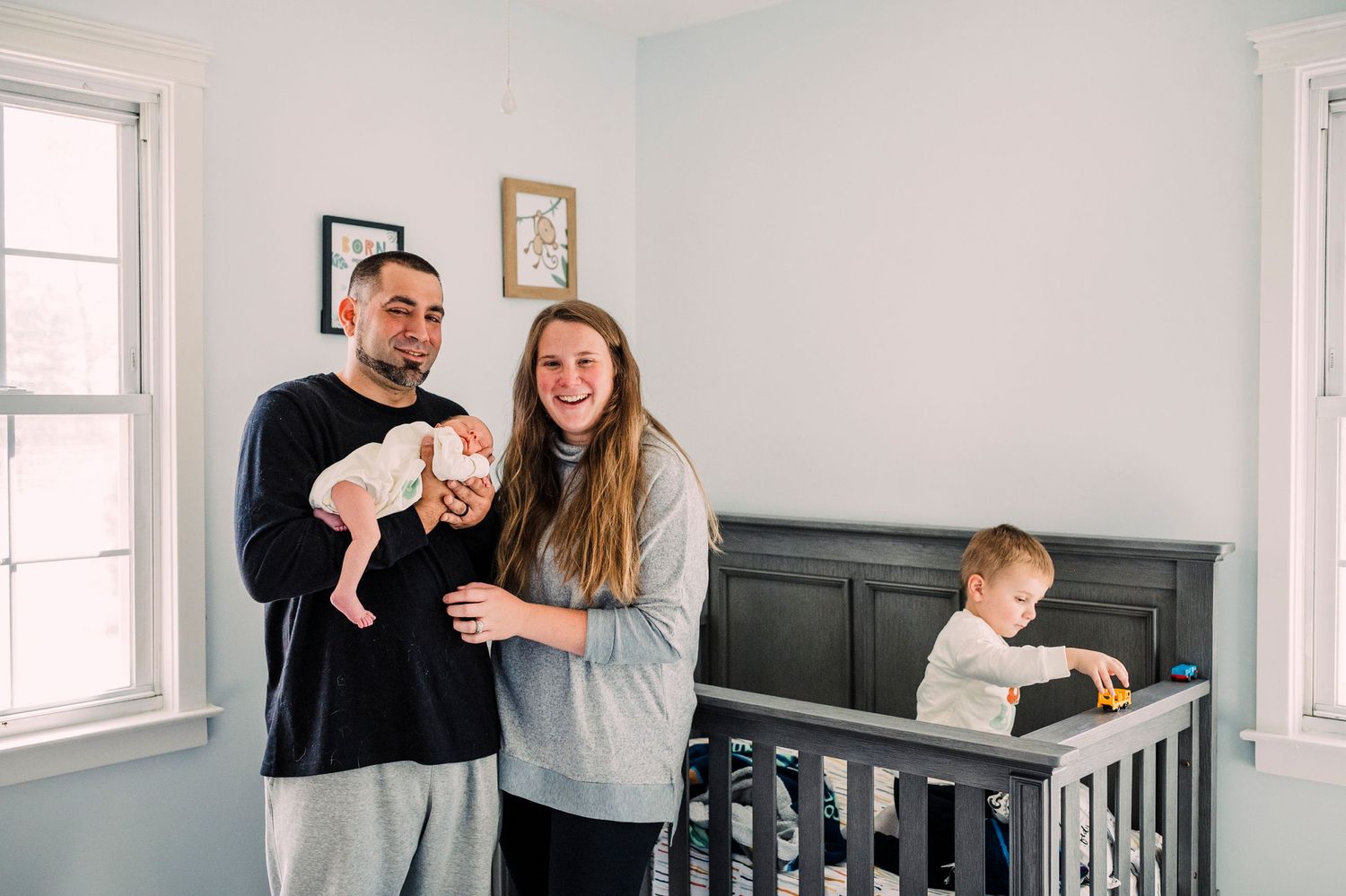 A family stands together in a bright bedroom with light walls and windows while a toddler sits in a crib.