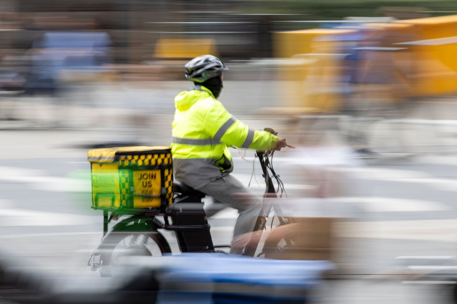 Motion-blurred cyclist wearing a high-vis jacket rides a delivery bike with a crate through a busy urban street.