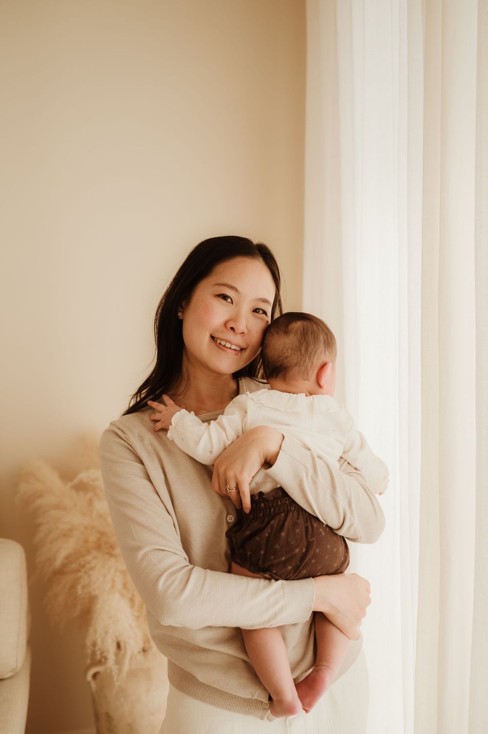A tender moment captured by window light as a baby in white is held in loving embrace against a soft beige background.