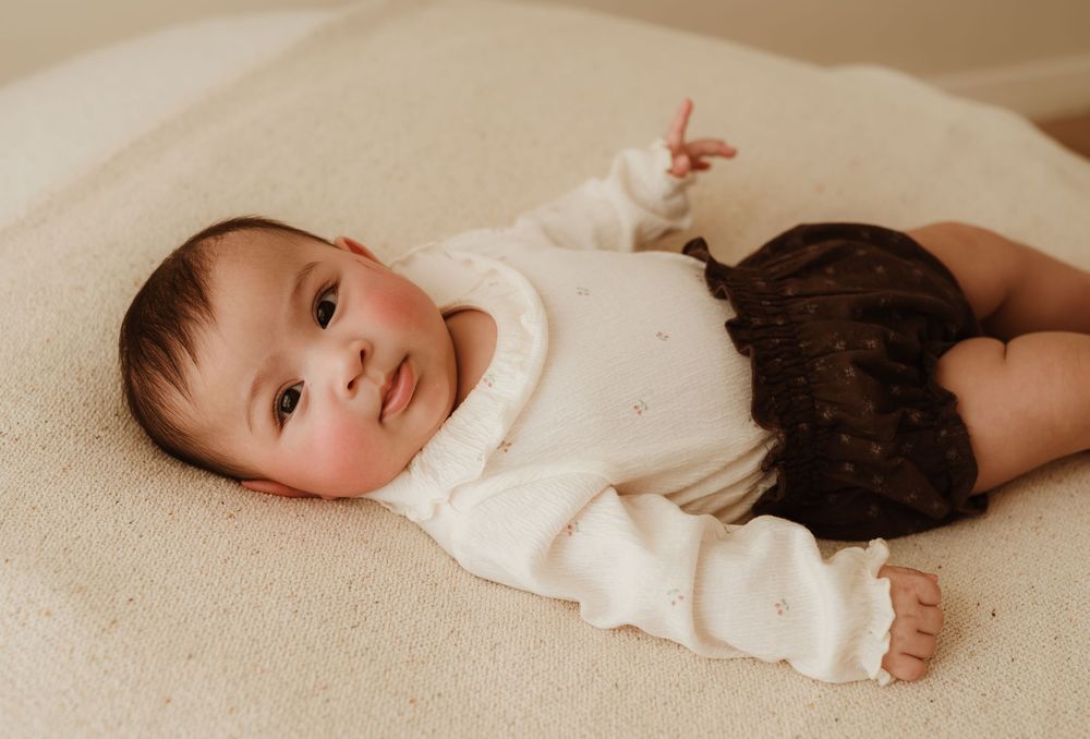 Baby wearing cream sweater and dark bloomers lies on soft beige surface in natural light setting.