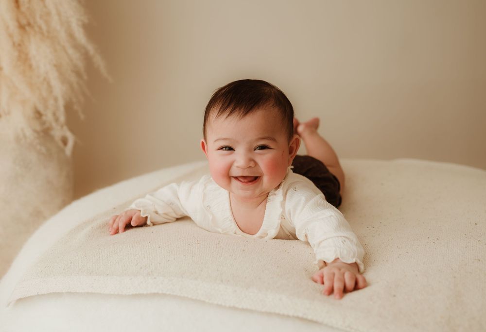 Small baby wearing white sweater lying on cream-colored textured surface with joyful expression.