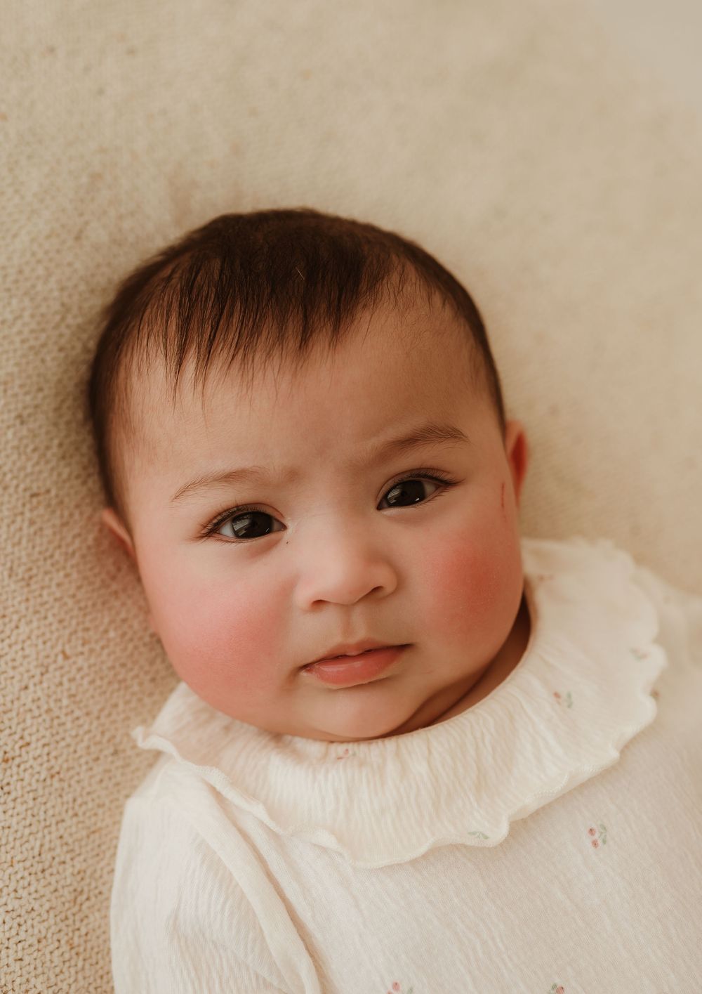 Sweet baby wearing a white ruffled collar smiles softly while laying on a beige background.