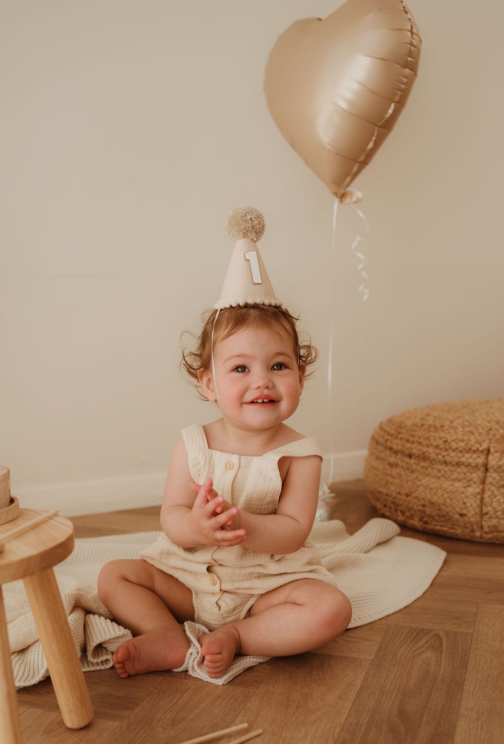 Baby wearing party hat sits on wooden floor with heart-shaped balloon and decorative elements in warm neutral tones.