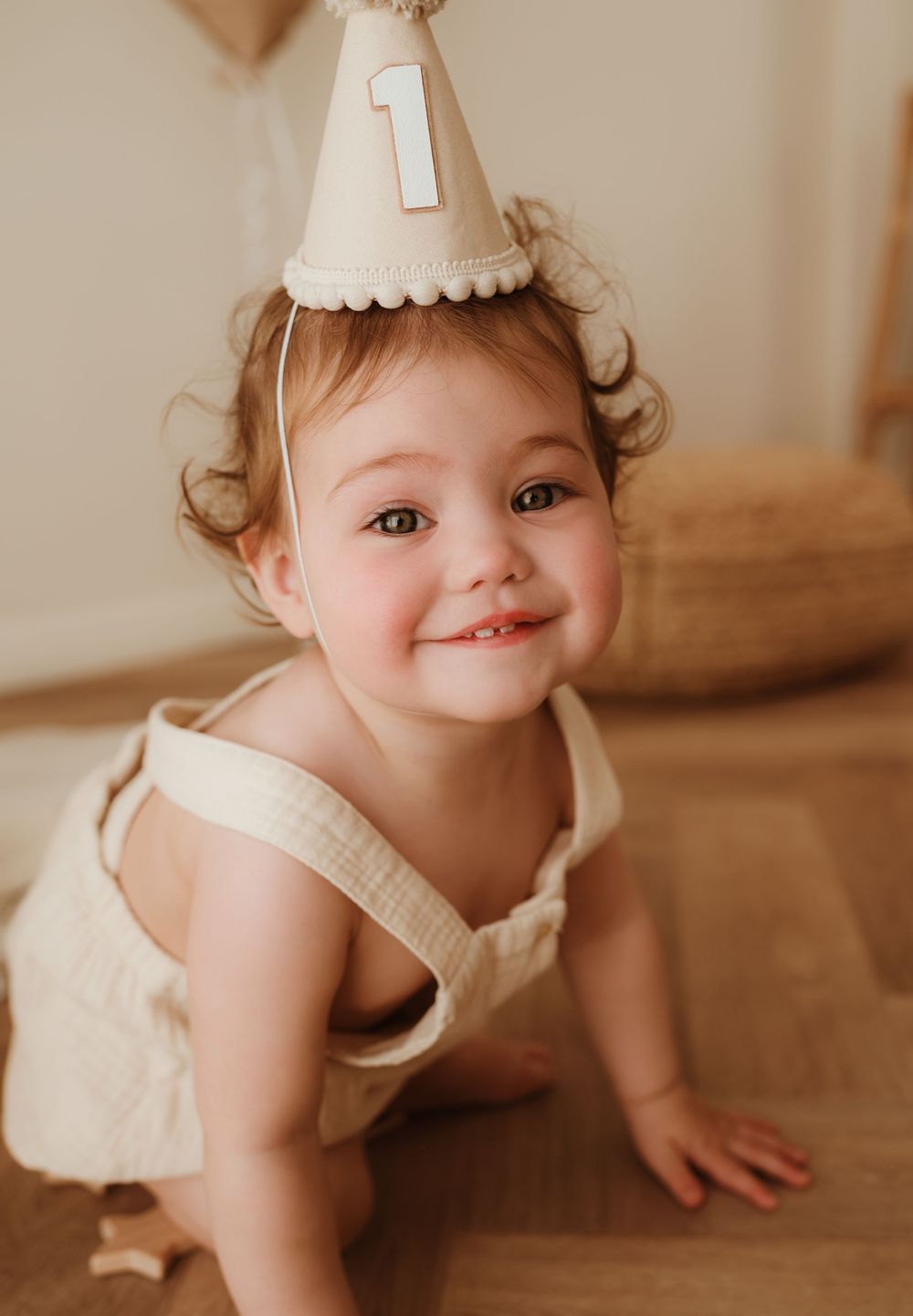 A toddler wearing a white dress and party hat with number 1 crawls on a wooden floor in warm lighting.