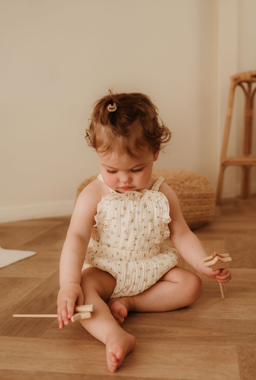 Baby in cream-colored romper sits on hardwood floor playing with wooden sticks in warm natural lighting.