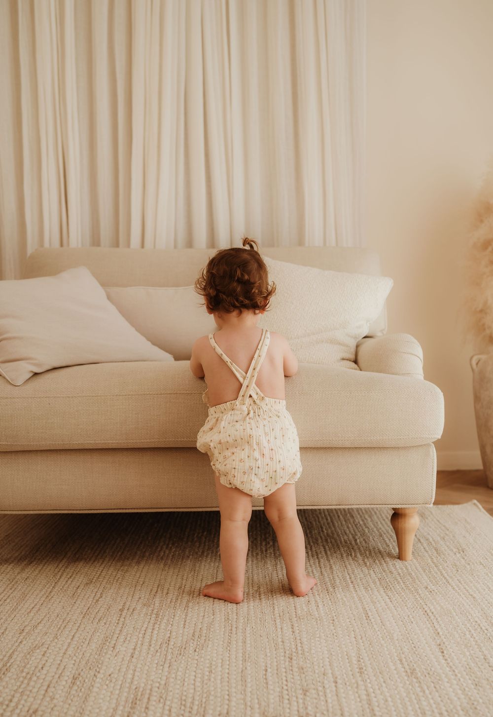 A toddler in a floral romper stands facing a cream-colored couch in a minimalist living room setting.
